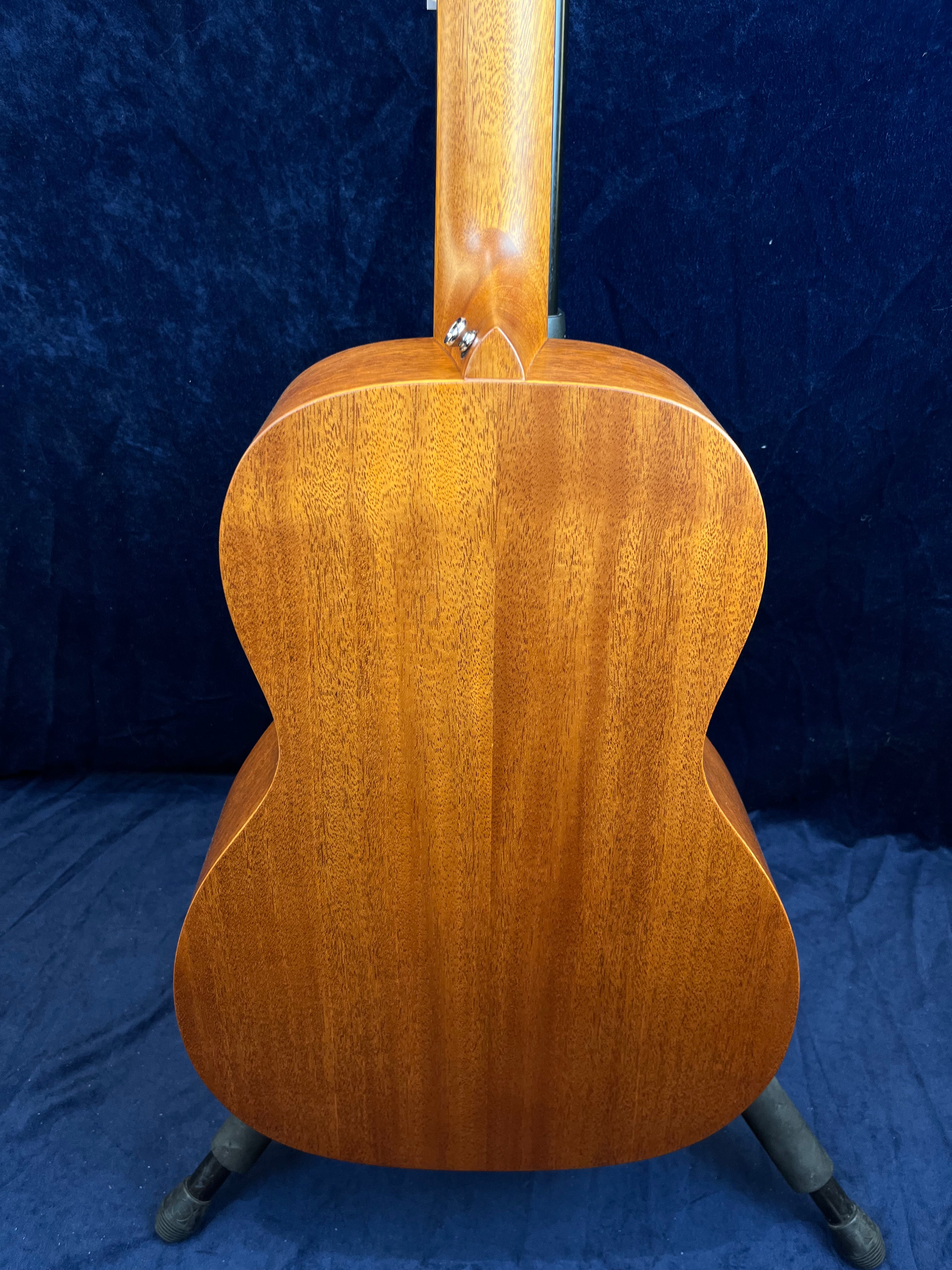 The back of an acoustic guitar made of smooth, rich brown wood with a subtle grain pattern, shown standing upright against a dark background.