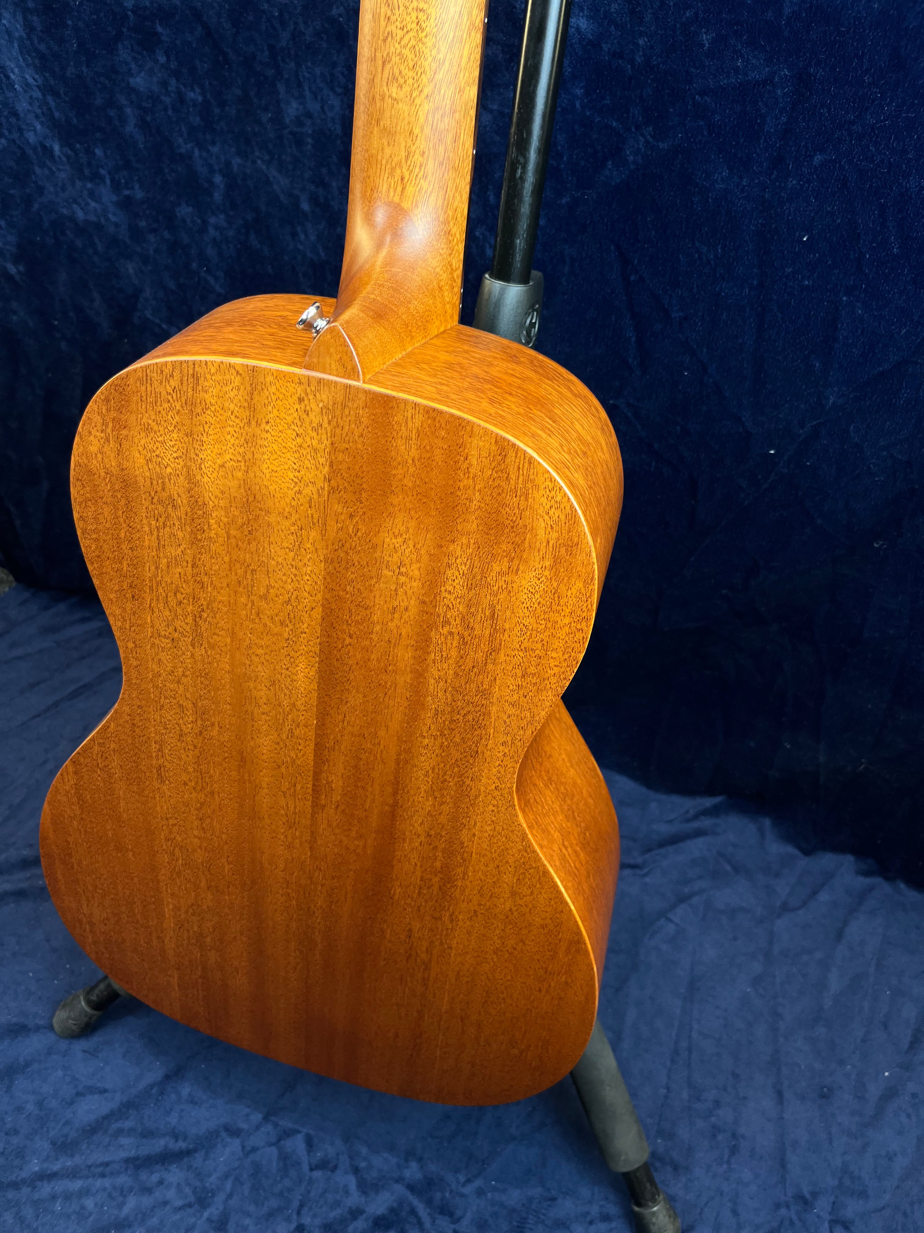 Acoustic guitar on a stand, showing the back and neck made of smooth, medium brown wood with a natural grain pattern.