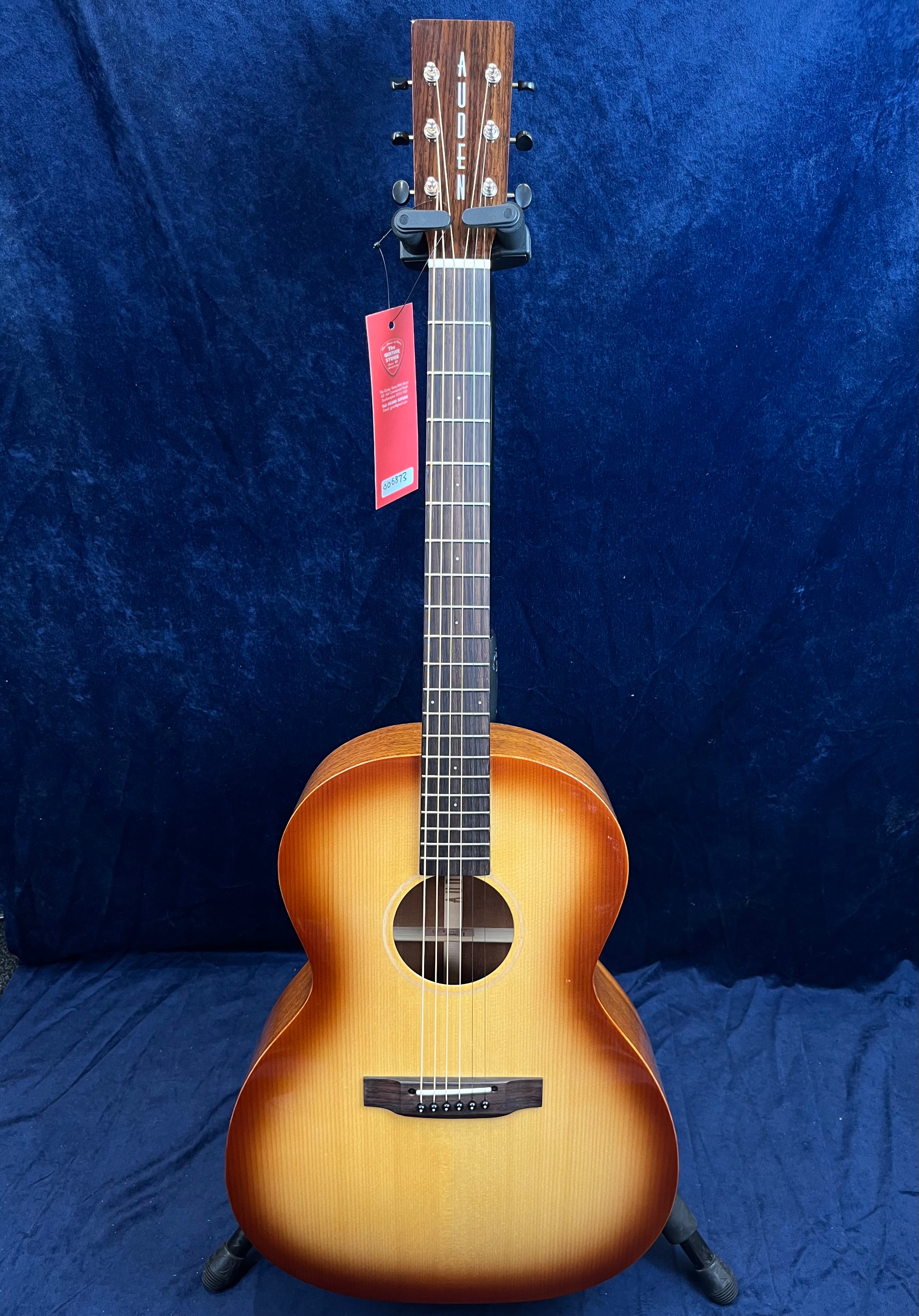 Guitare acoustique couleur sunburst dégradé du jaune au brun, forme arrondie, tête avec inscription Auden, étiquette rouge accrochée au manche.