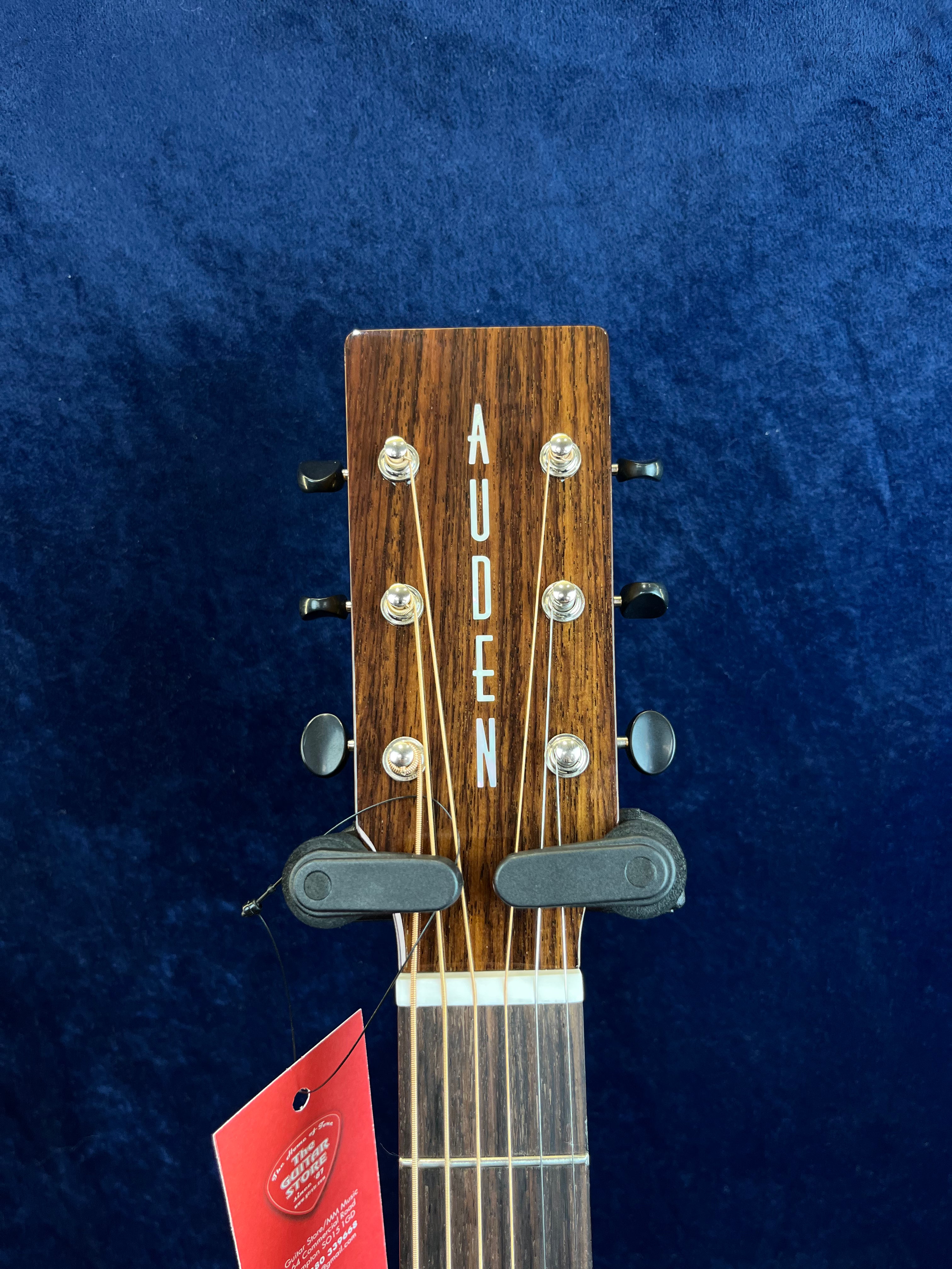 Tête d’une guitare acoustique en bois foncé avec le nom AUDEN en lettres blanches et deux capos noirs placés sur le haut du manche.