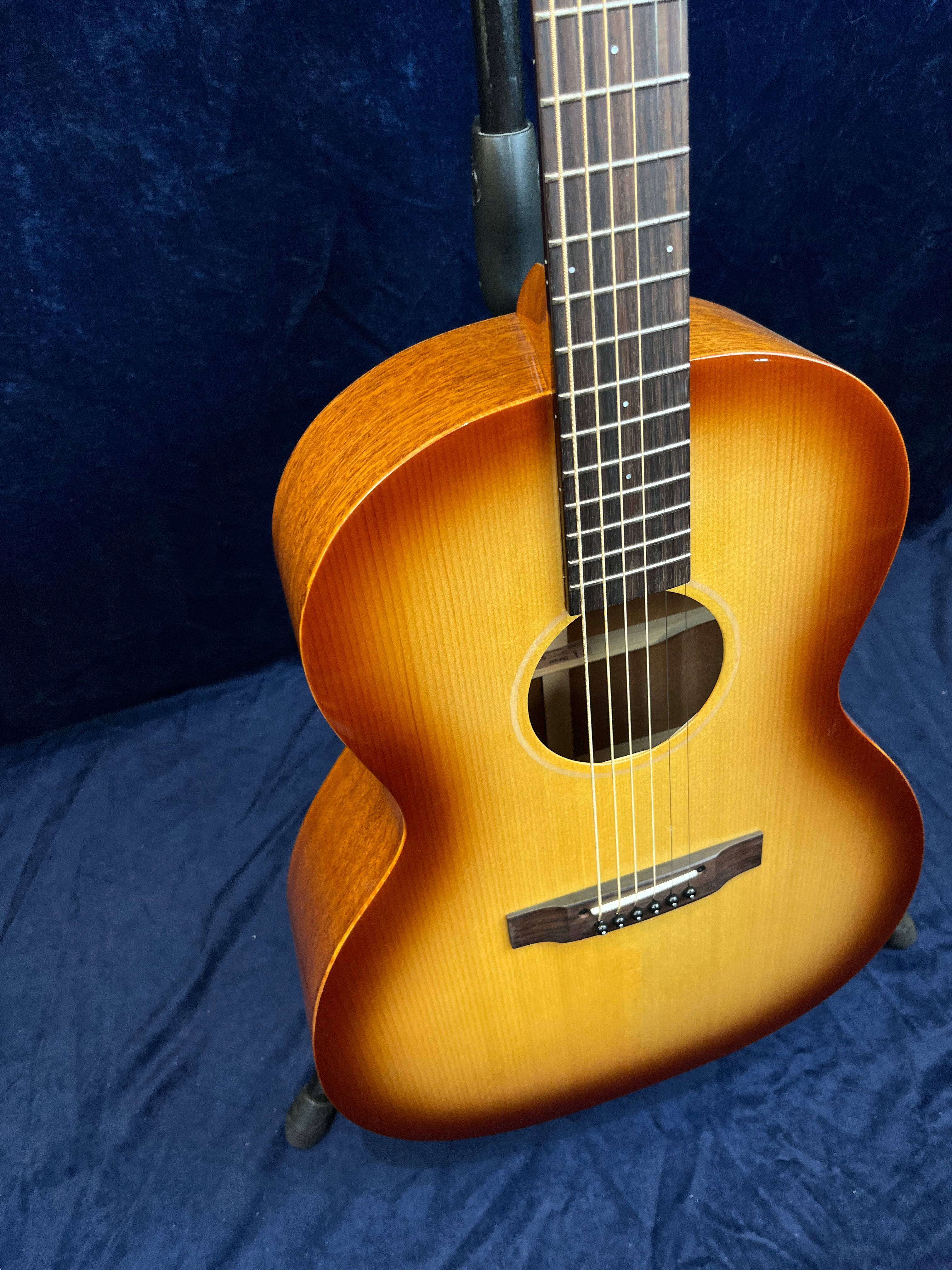 Guitare acoustique à table bombée, finition dégradée du jaune au brun, six cordes, touche sombre, posée sur un fond bleu.