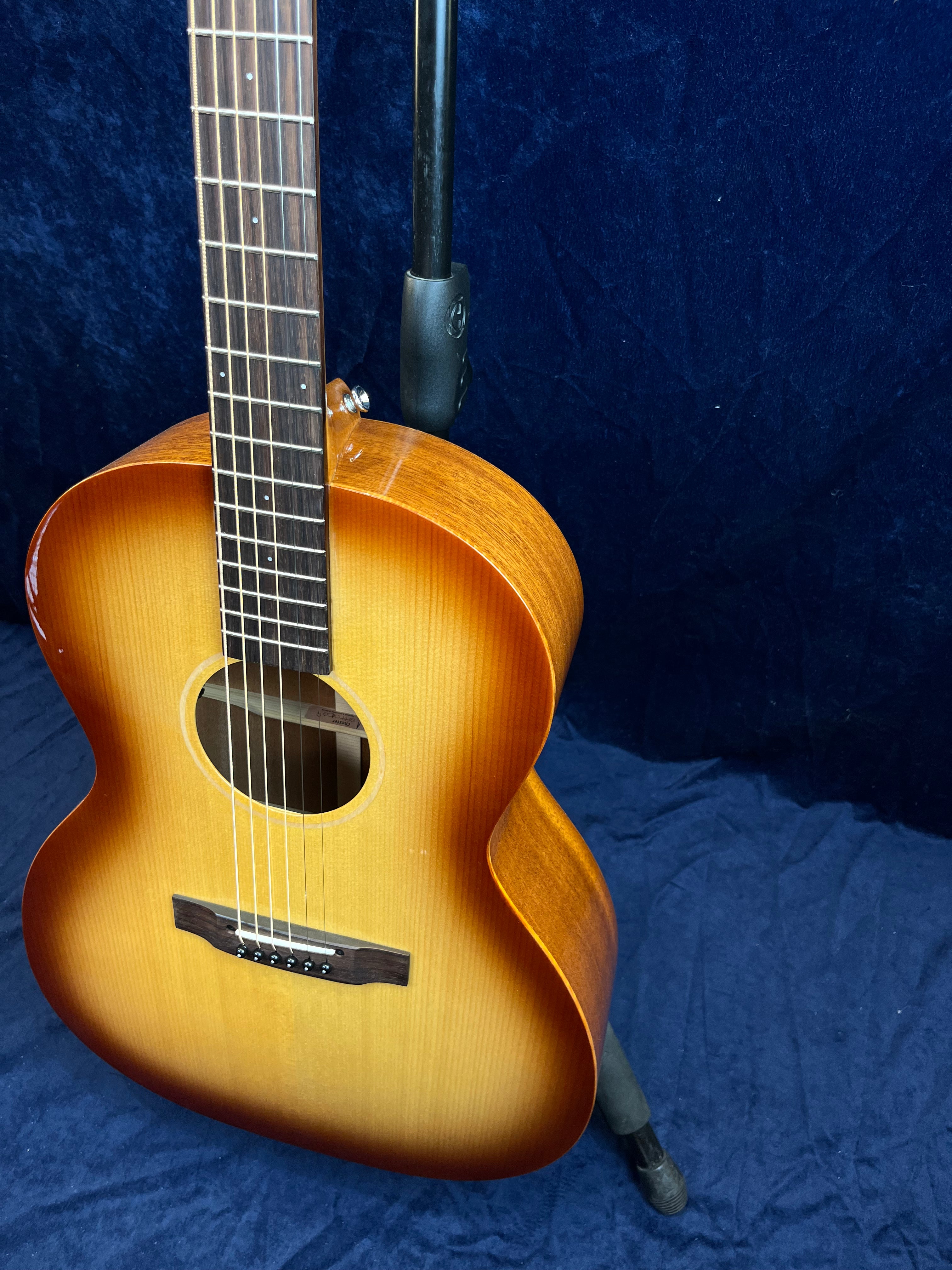Guitare acoustique posée sur un support, caisse en bois vernis avec dégradé brun clair à foncé, six cordes et touche en bois foncé.