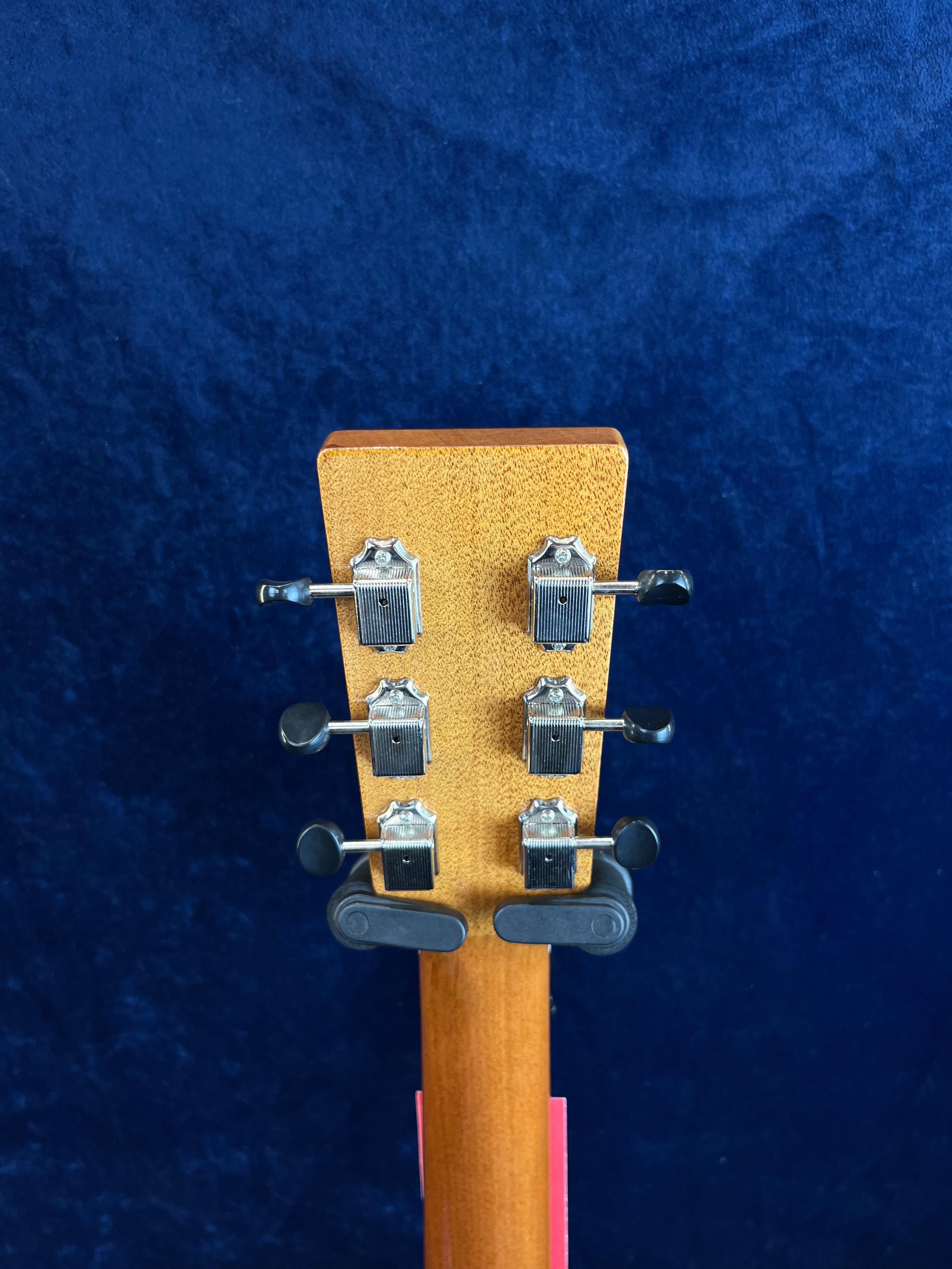 Tête de guitare acoustique en bois avec six mécaniques argentées alignées par trois de chaque côté, fond bleu uni.