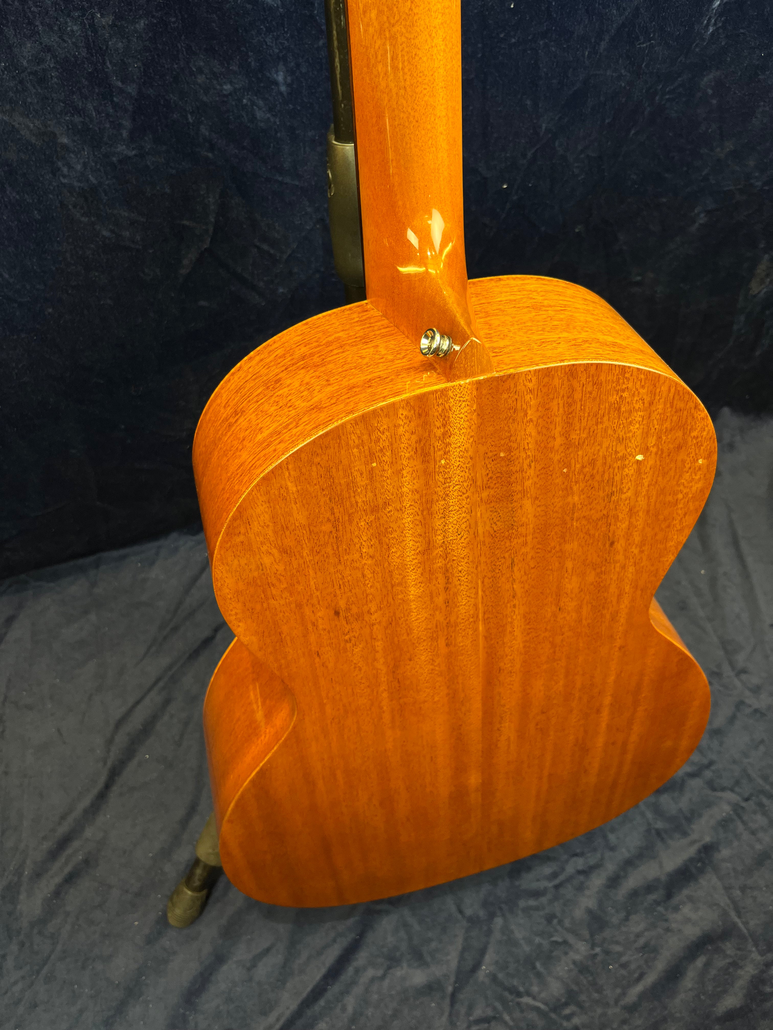 Dos d’une guitare acoustique en bois brun clair au grain visible, fixée par une attache métallique proche du manche, fond noir en arrière-plan.
