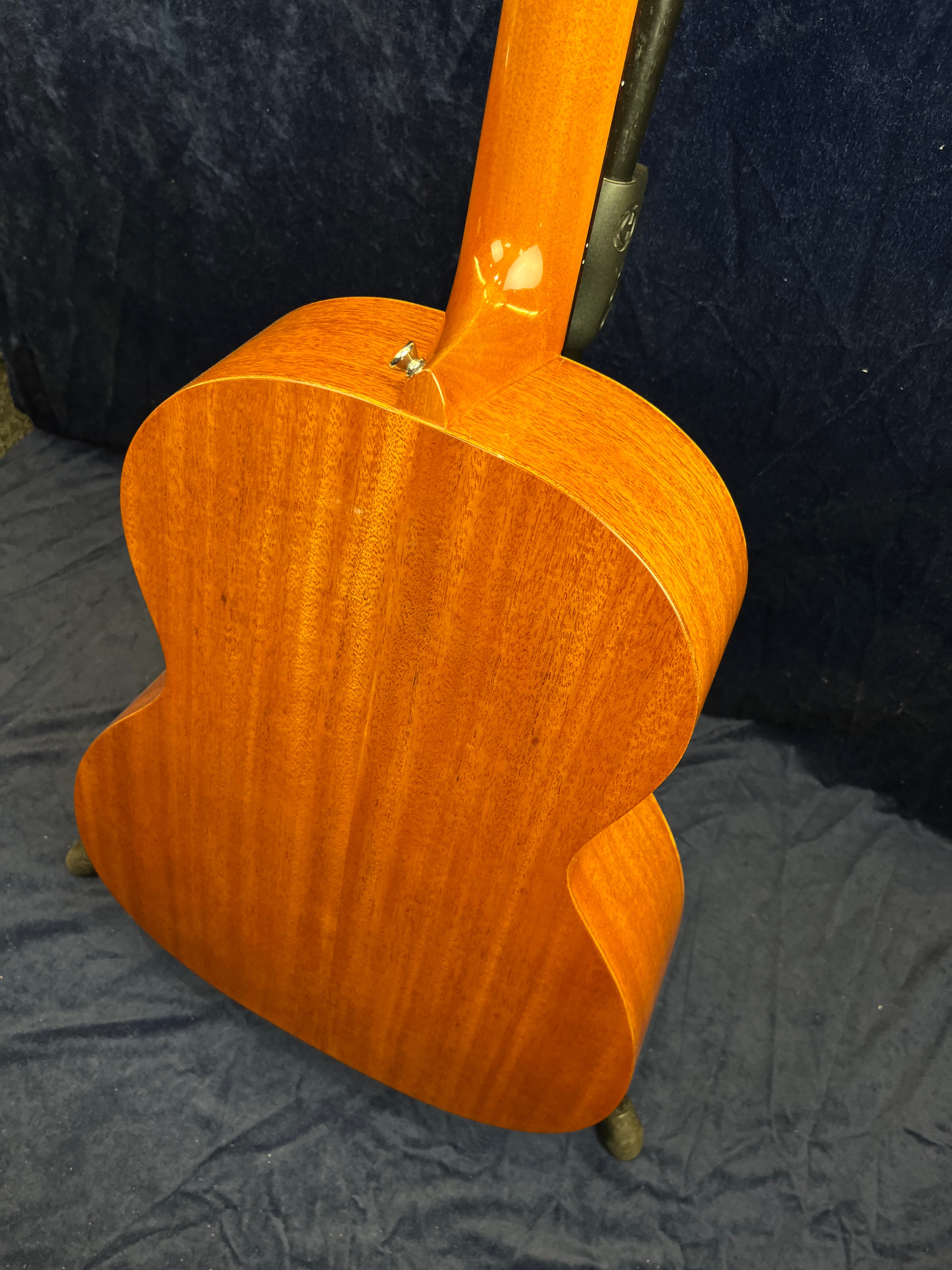 Guitare acoustique vue de dos, bois verni couleur miel à grain visible, posée sur un support, fond en tissu foncé.