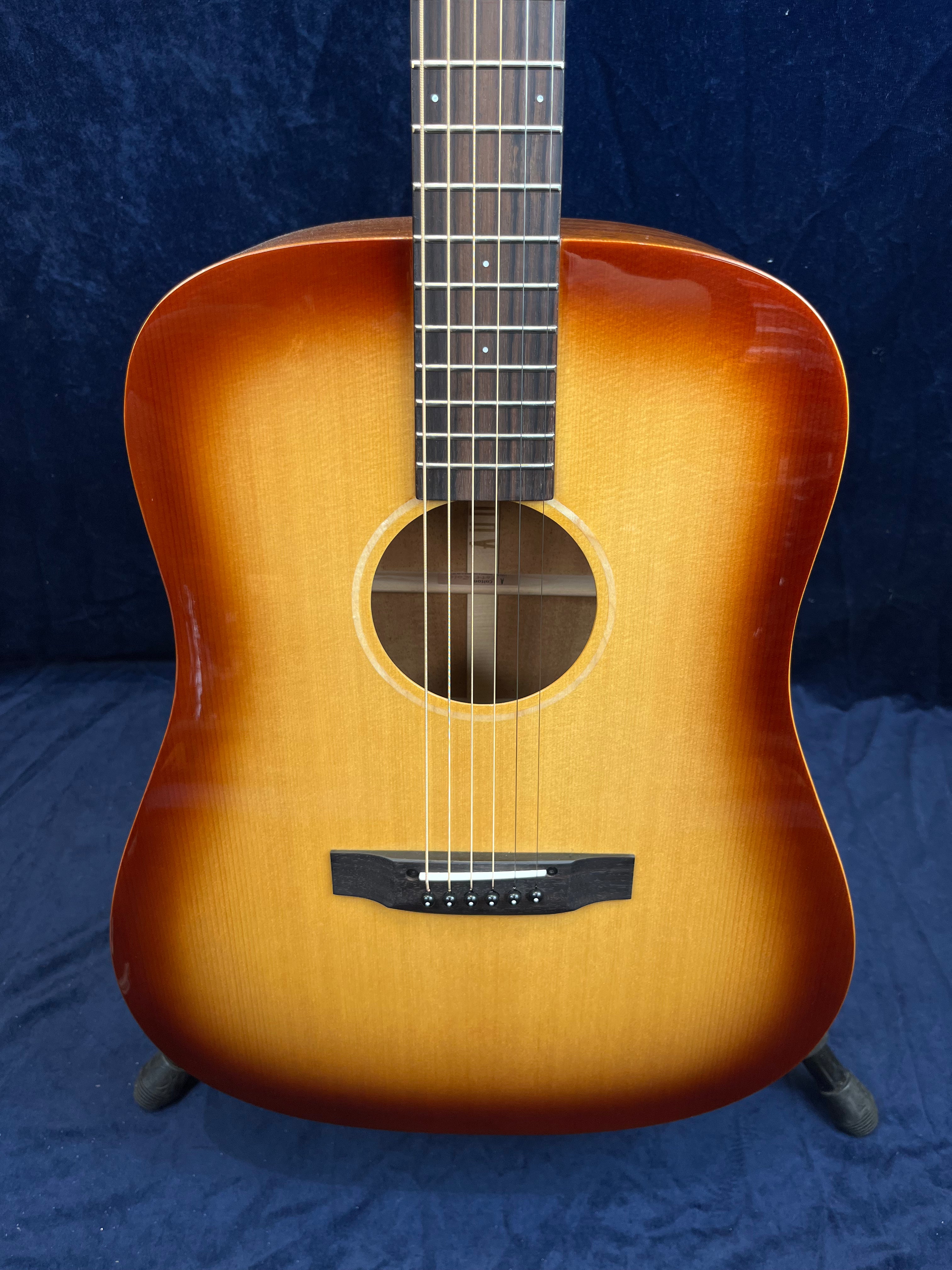 Acoustic guitar with a sunburst finish, six strings, a round soundhole, and a dark wooden bridge, displayed upright against a dark background.