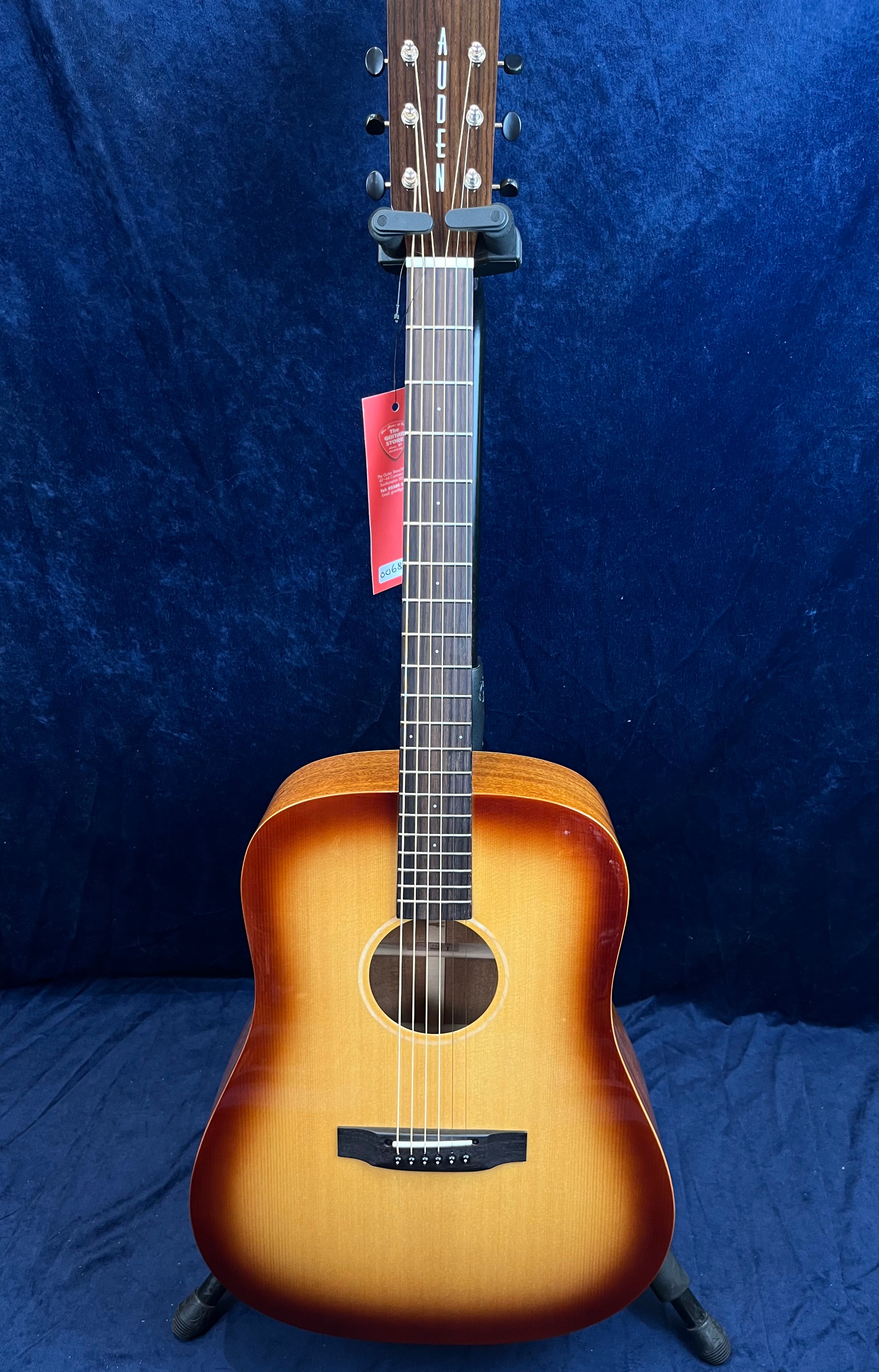 Auden acoustic guitar with a sunburst finish, six steel strings, wooden fretboard, and a red tag hanging from the neck against a dark blue background.