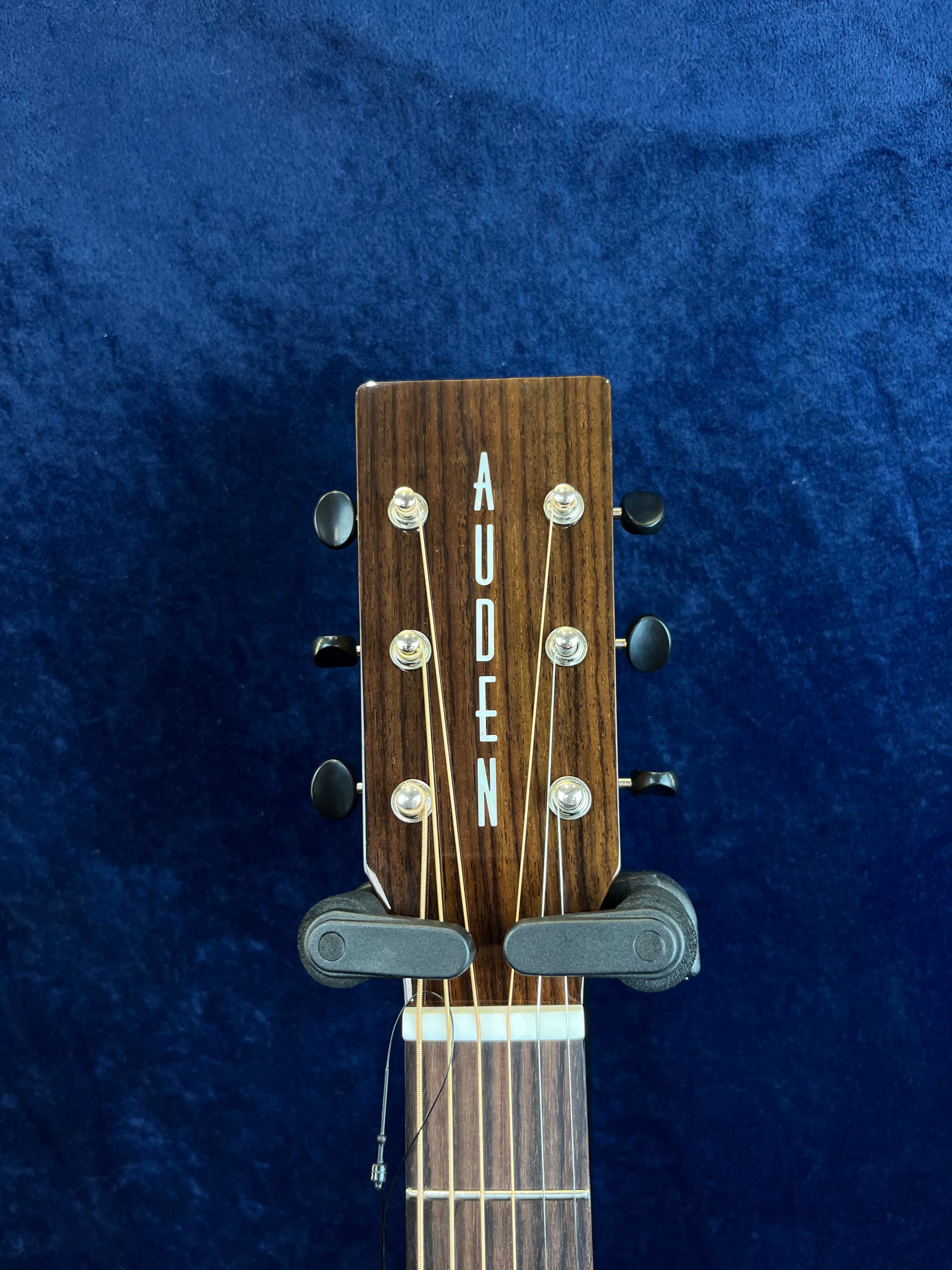 Wooden headstock of an acoustic guitar with "AUDEN" in white letters, six tuning pegs, and strings, set against a dark blue background.