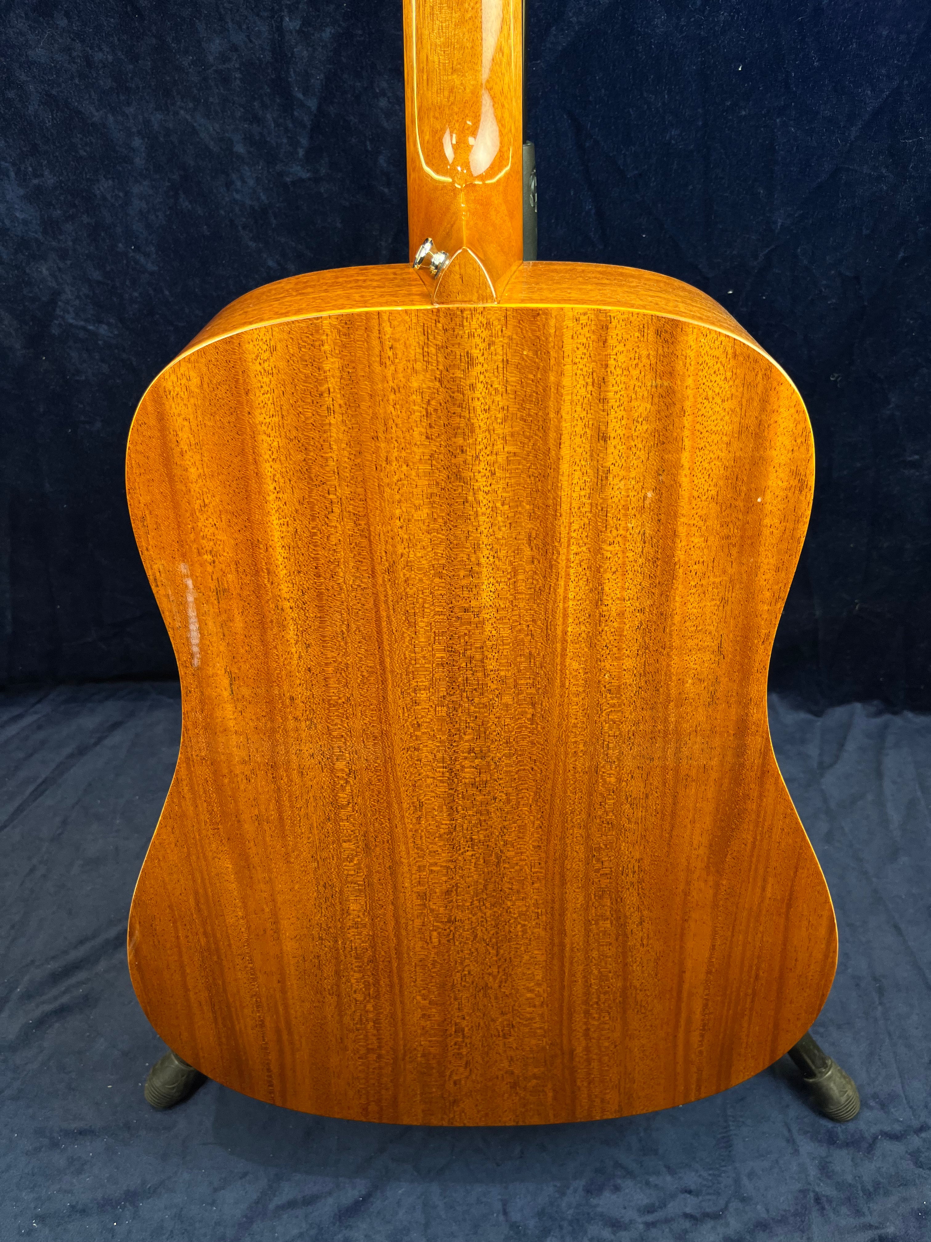 Glossy mahogany back of an acoustic guitar with visible wood grain, rounded shoulders, and neck joint detail, set against a dark background.