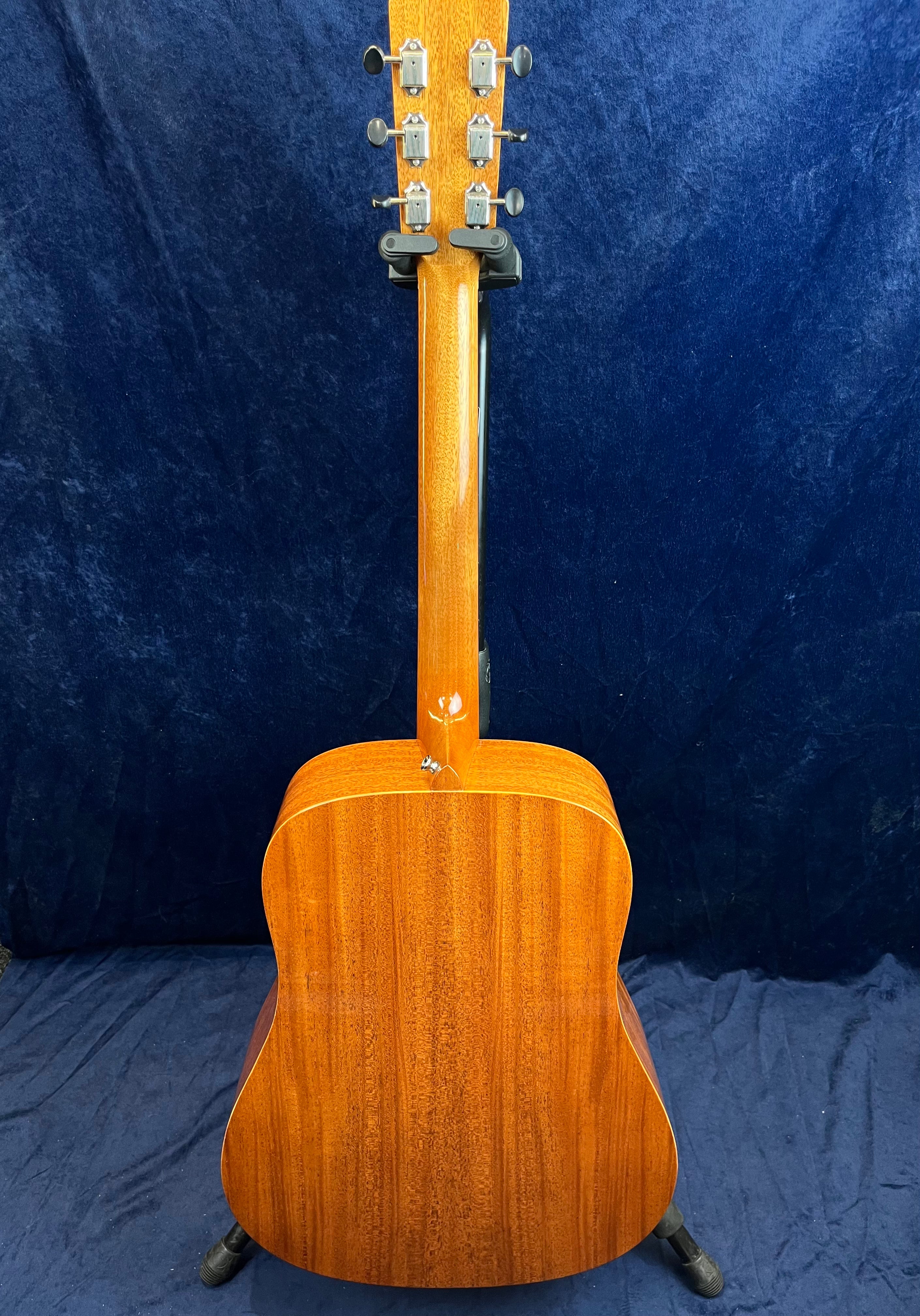 The back view of a wooden acoustic guitar with a glossy finish, showing tuning pegs, neck, and rich brown wood grain body against a dark blue background.