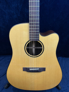 Light wood acoustic guitar with a cutaway body, six strings, round sound hole, dark bridge, and fretboard, shown upright against a dark background.