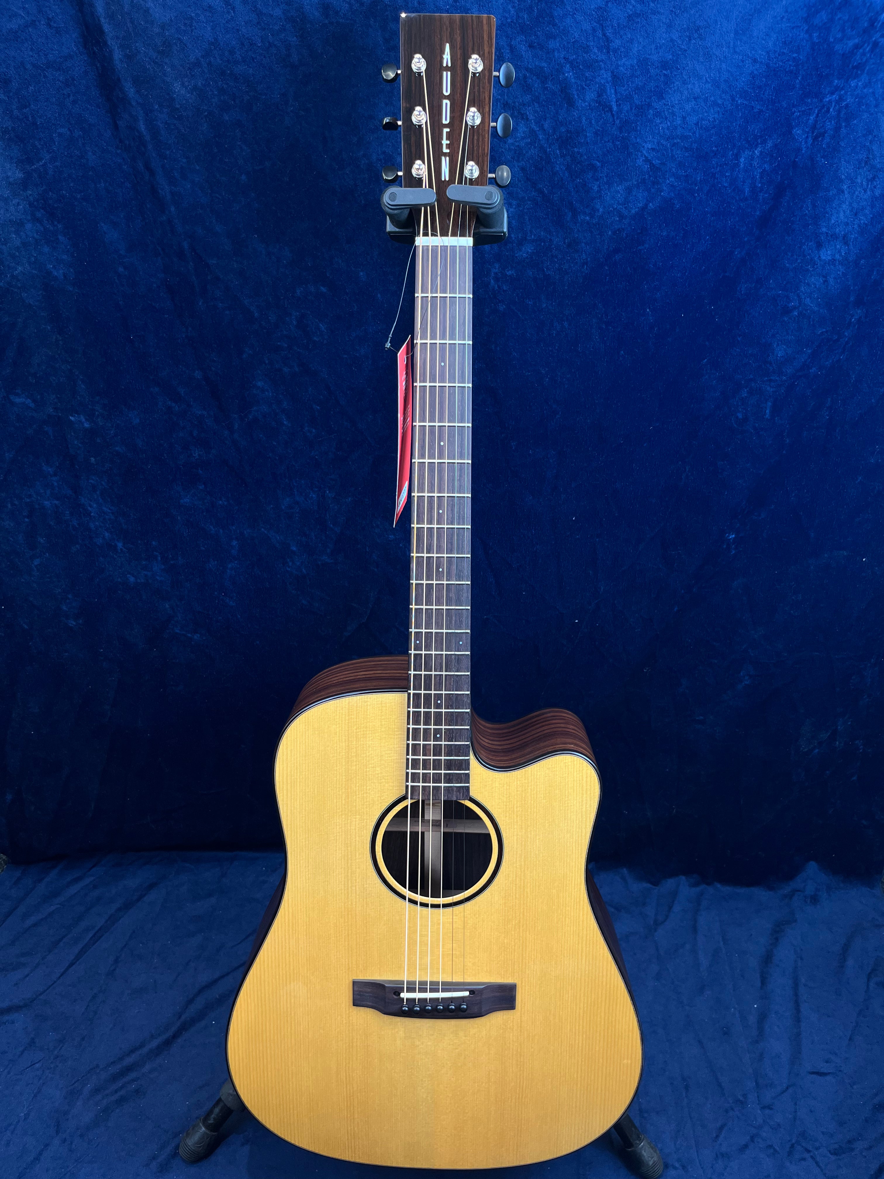 Guitare acoustique couleur bois clair avec pan coupé, six cordes, touche sombre et tête marquée du nom Auden, posée debout sur un support.