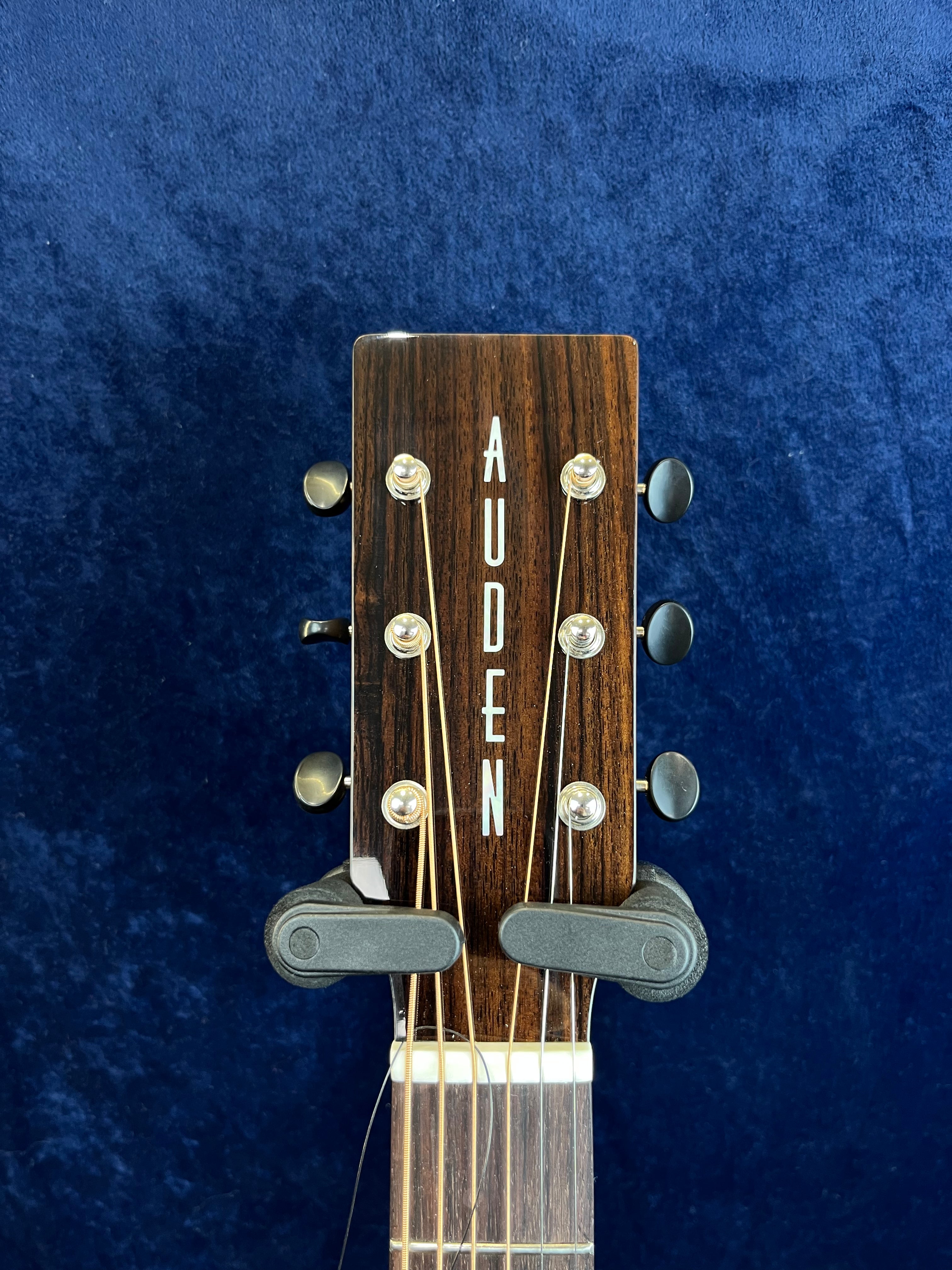 Tête de guitare acoustique en bois foncé avec le mot AUDEN en lettres verticales blanches, six mécaniques noires et un capodastre fixé.