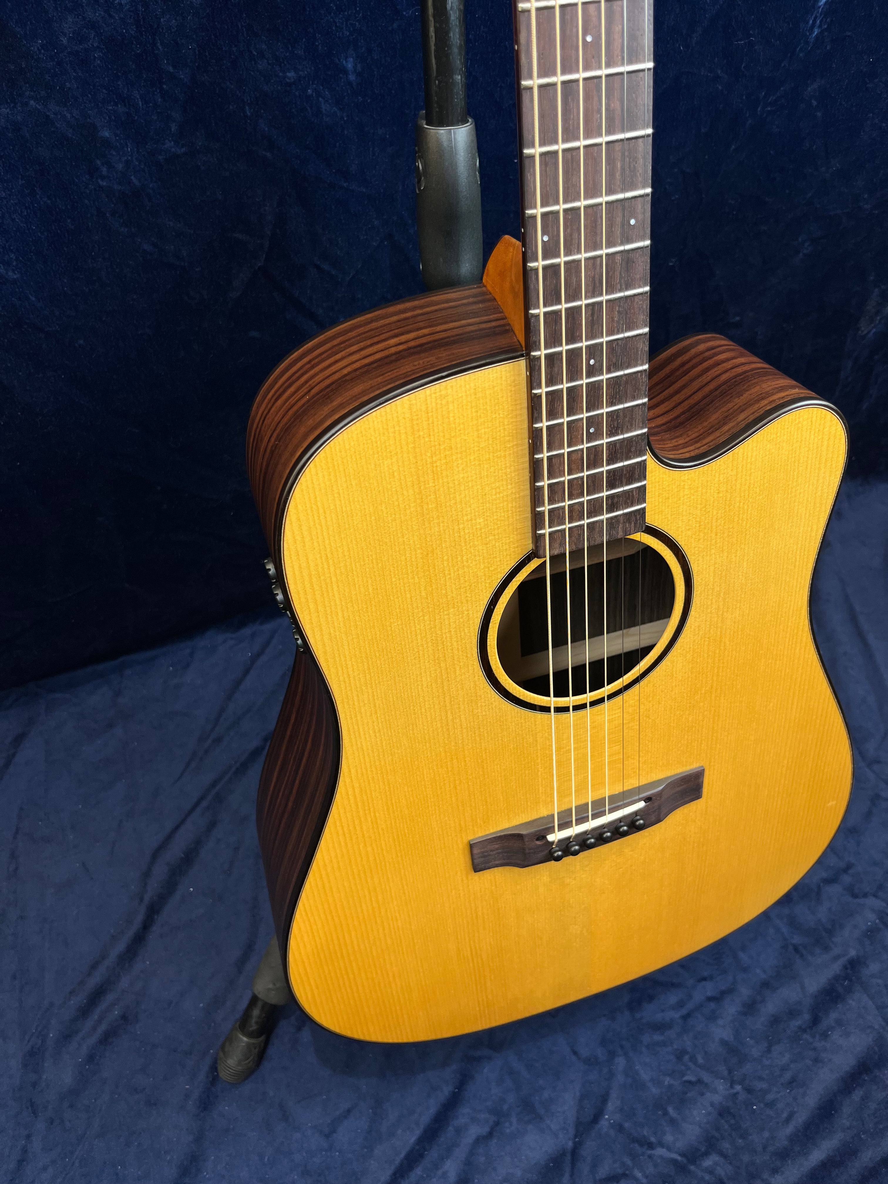 Guitare acoustique avec table en bois clair, éclisses et dos foncés à motif bois, pan coupé, posée sur un support contre un fond bleu.
