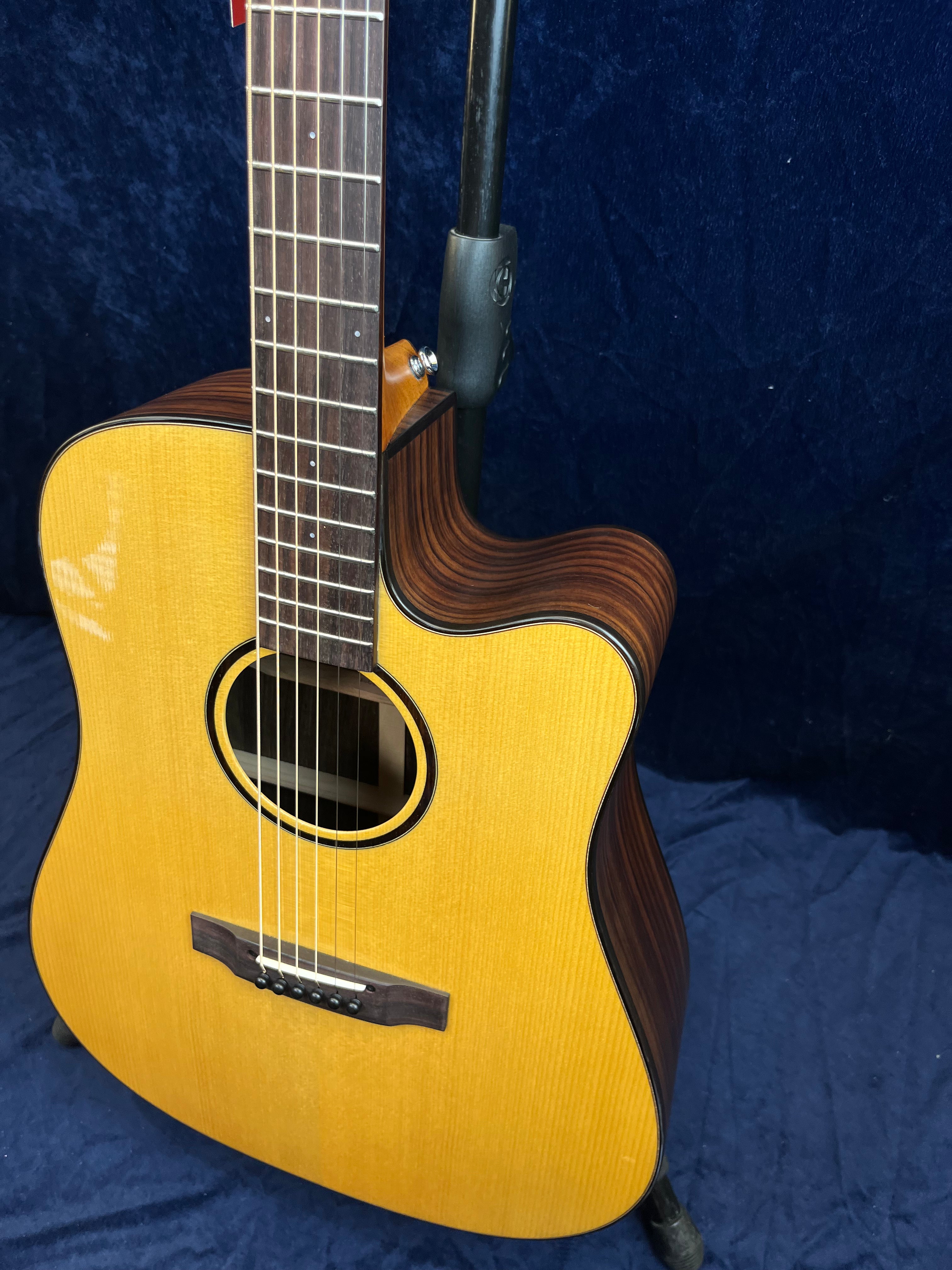 Guitare acoustique à pan coupé avec table en bois clair, éclisses foncées à rayures, six cordes, posée sur un support contre un fond bleu.
