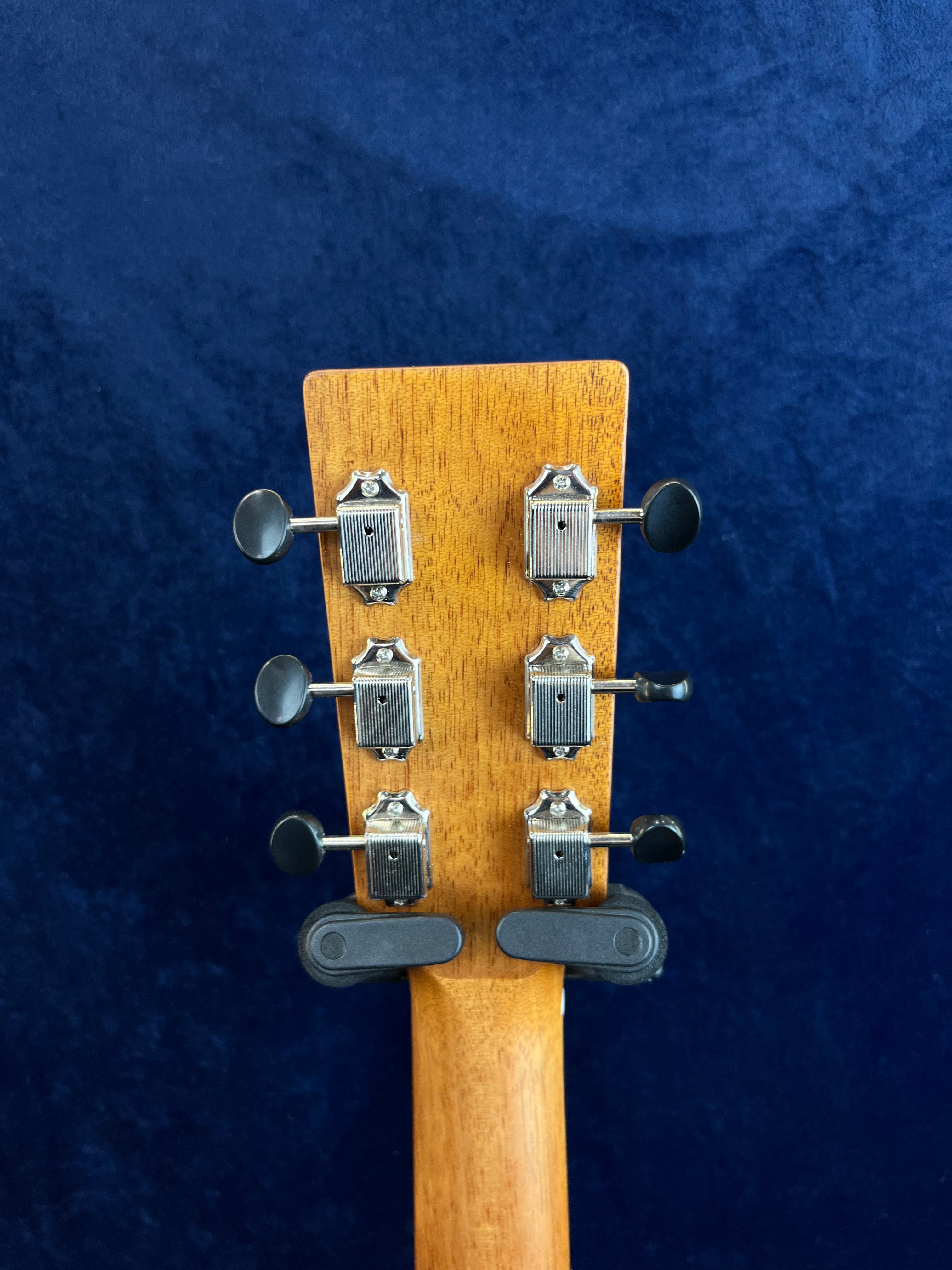Tête de guitare acoustique en bois clair, vue de dos, avec six mécaniques argentées alignées verticalement, fond bleu foncé uniforme.
