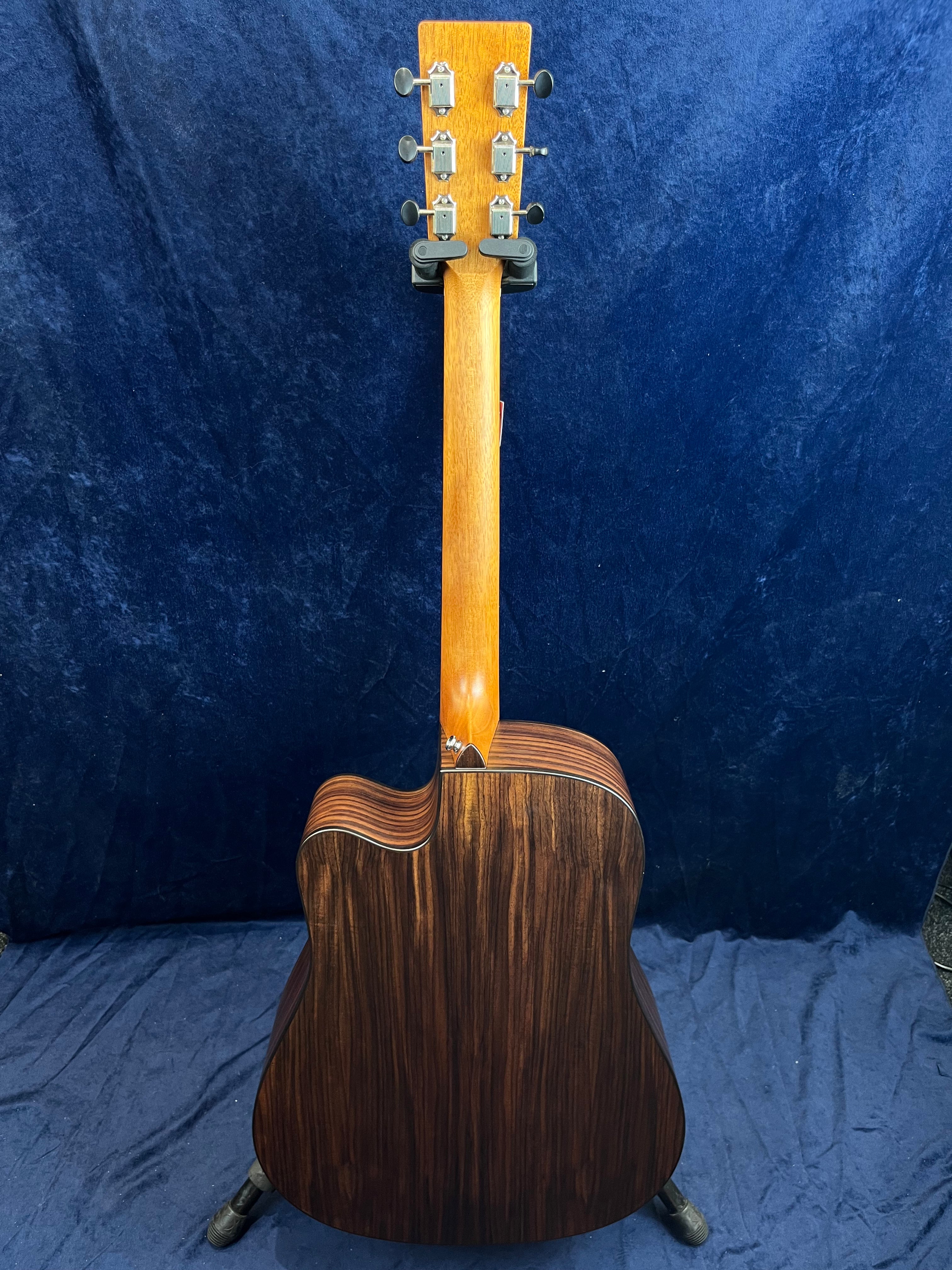 Guitare acoustique vue de dos, caisse en bois foncé nervuré, manche clair, mécaniques métalliques visibles, posée sur un support noir.