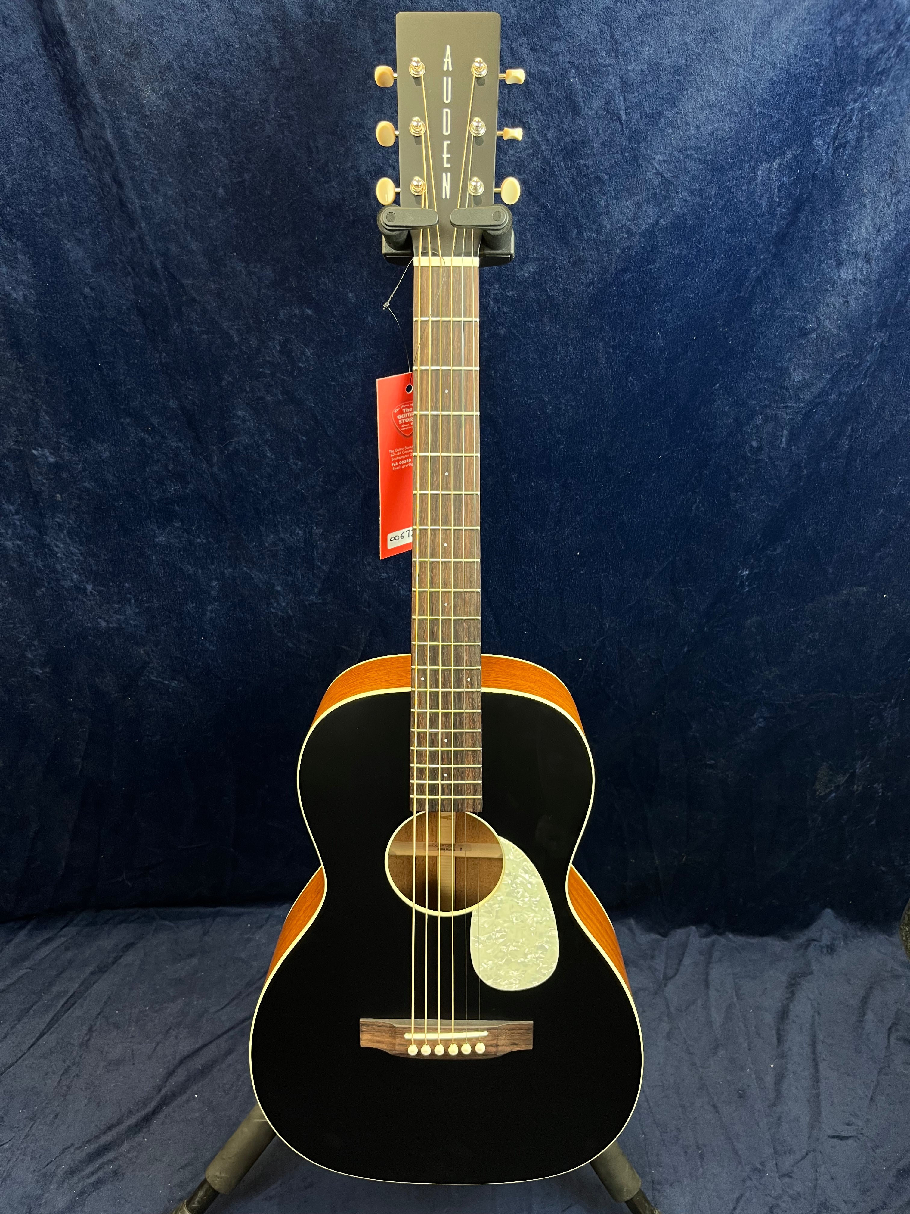 Black Auden acoustic guitar with a light wooden bridge, pearl pickguard, and visible red tag hanging from the headstock.