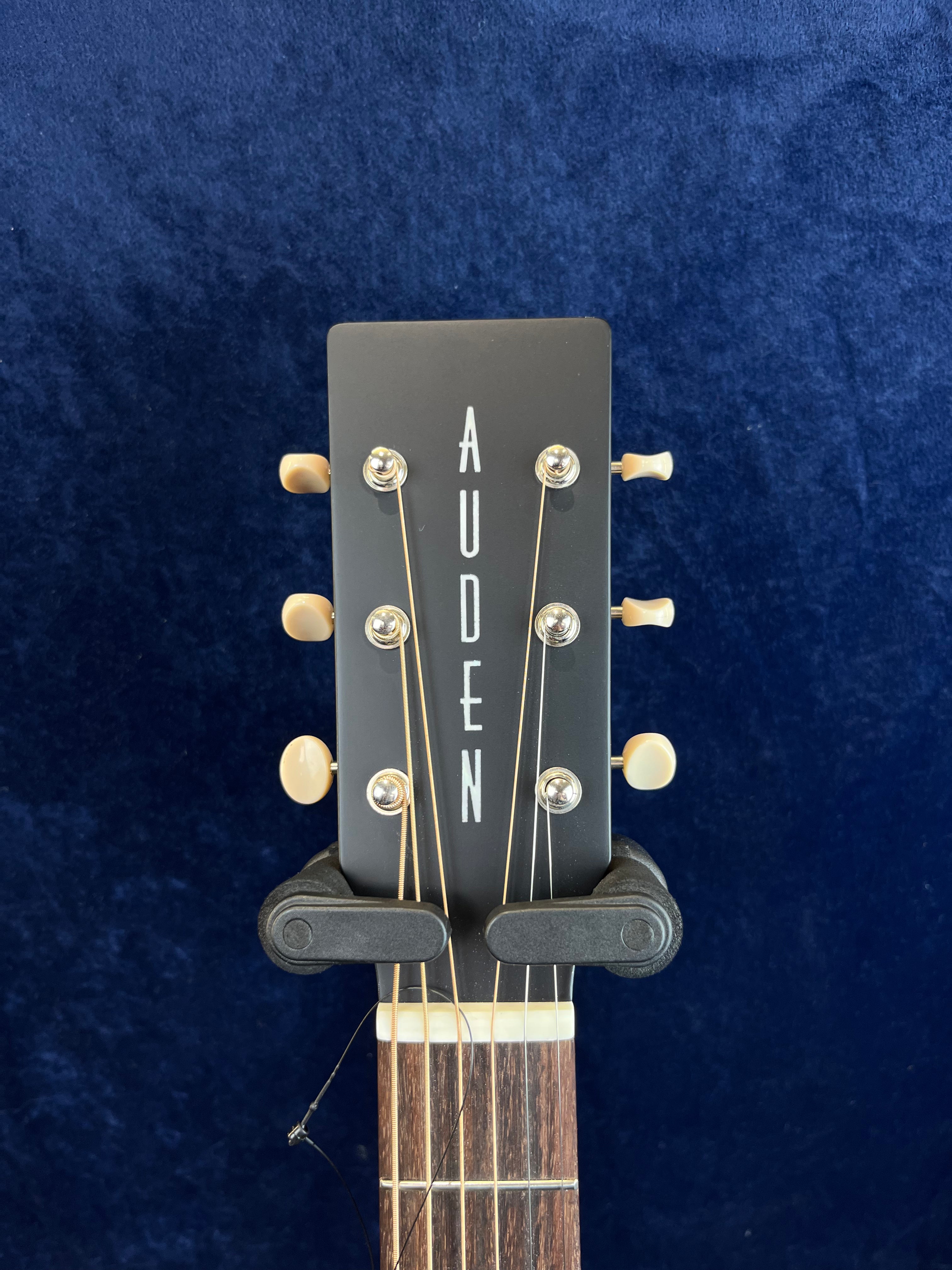 Black Auden acoustic guitar headstock with cream tuning pegs, vertical white "AUDEN" lettering, and six strings on a dark blue background.