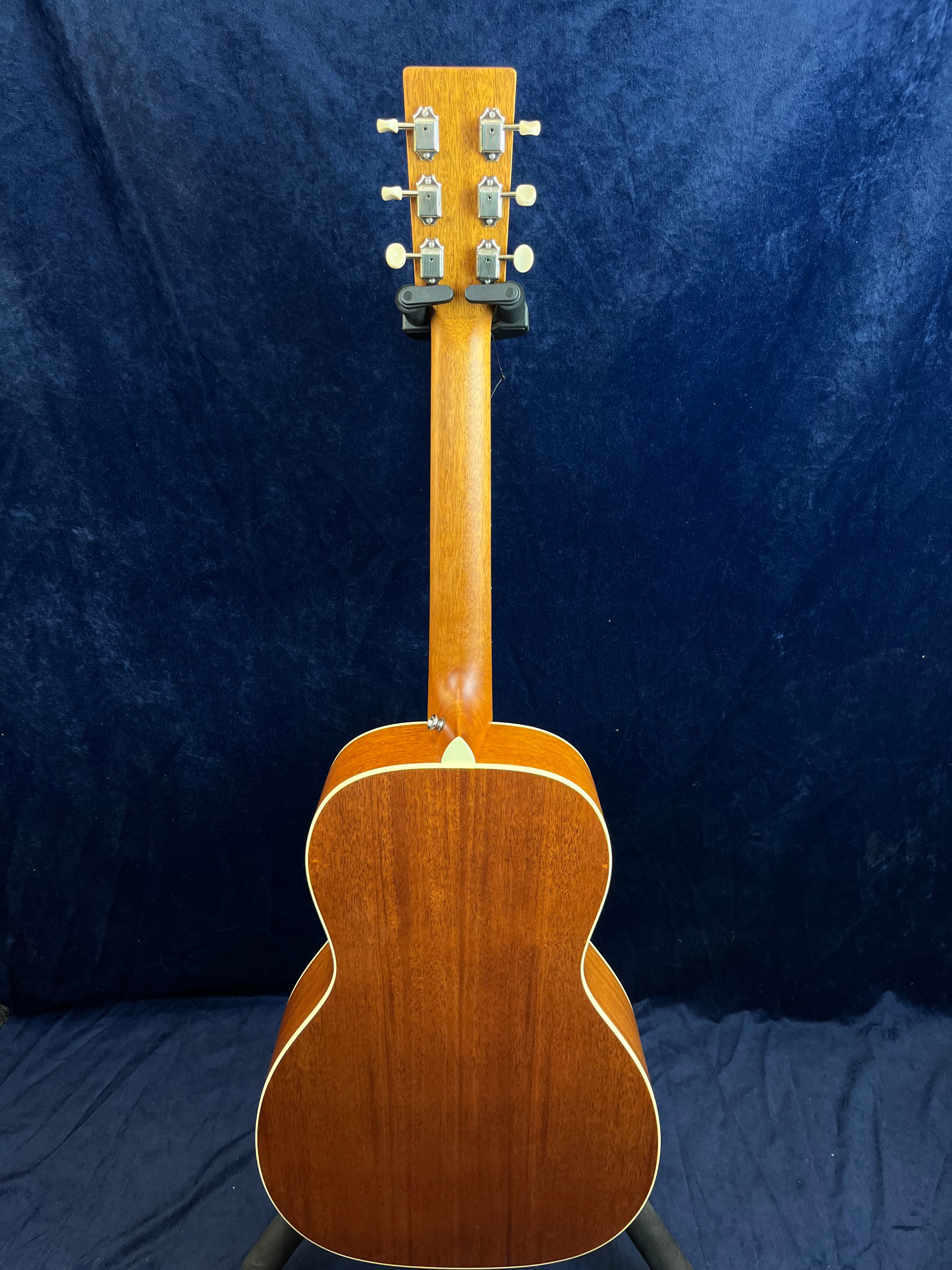 The back of an acoustic guitar with a mahogany finish, six tuning pegs, and cream edge binding is shown upright against a dark blue background.