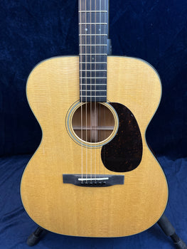 Acoustic guitar with light wood top, dark pickguard, six strings, and a round sound hole, shown standing upright against a dark background.