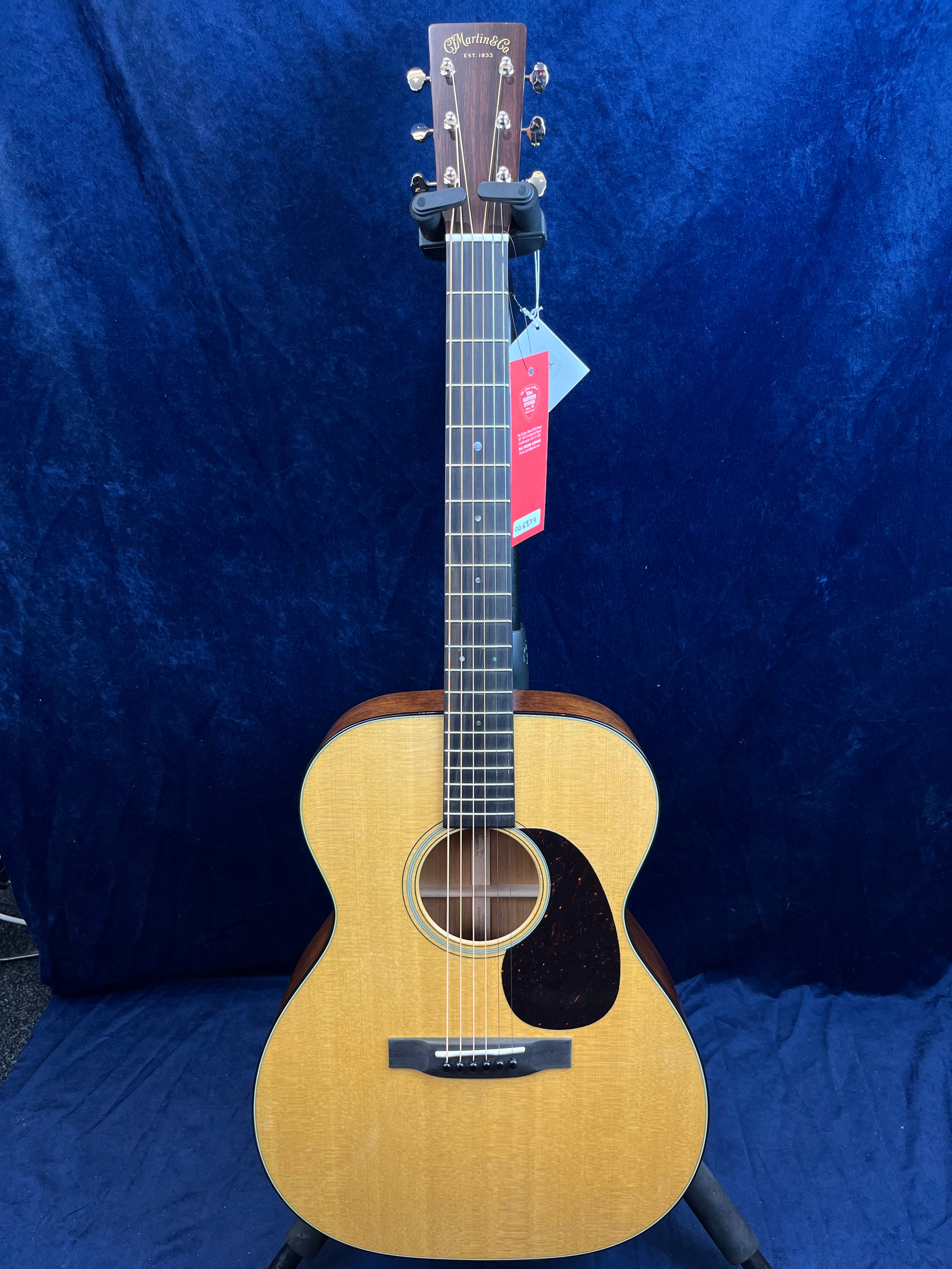 Light wood Martin acoustic guitar with a dark pickguard, six strings, and gold tuning pegs, displayed upright against a dark blue background.