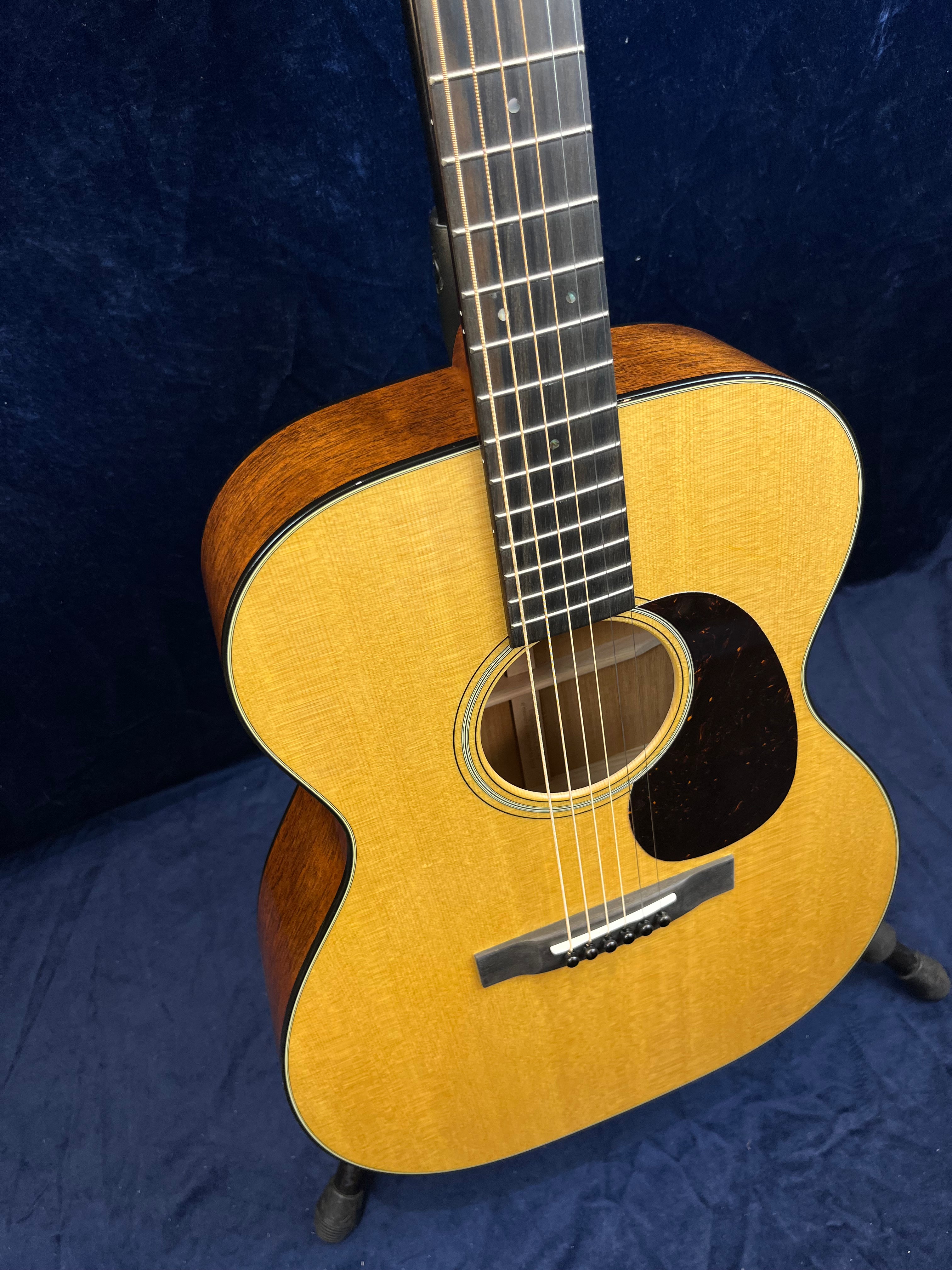 Acoustic guitar with a light wood top, dark wood sides, six strings, and a black pickguard, resting on a stand against a dark blue background.