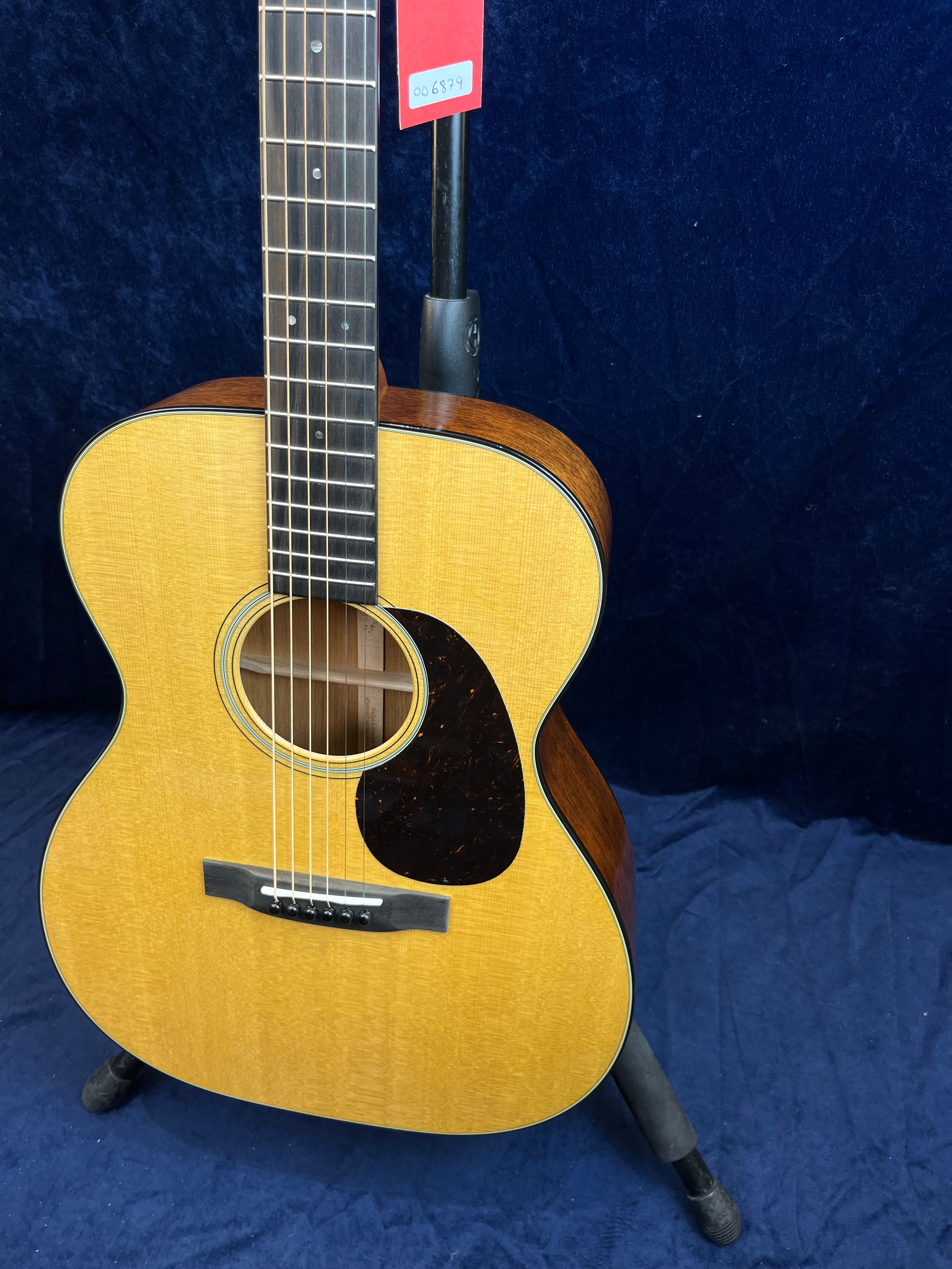 Light wood acoustic guitar with a dark pickguard, six strings, and a dark fingerboard, resting on a stand against a dark blue background.