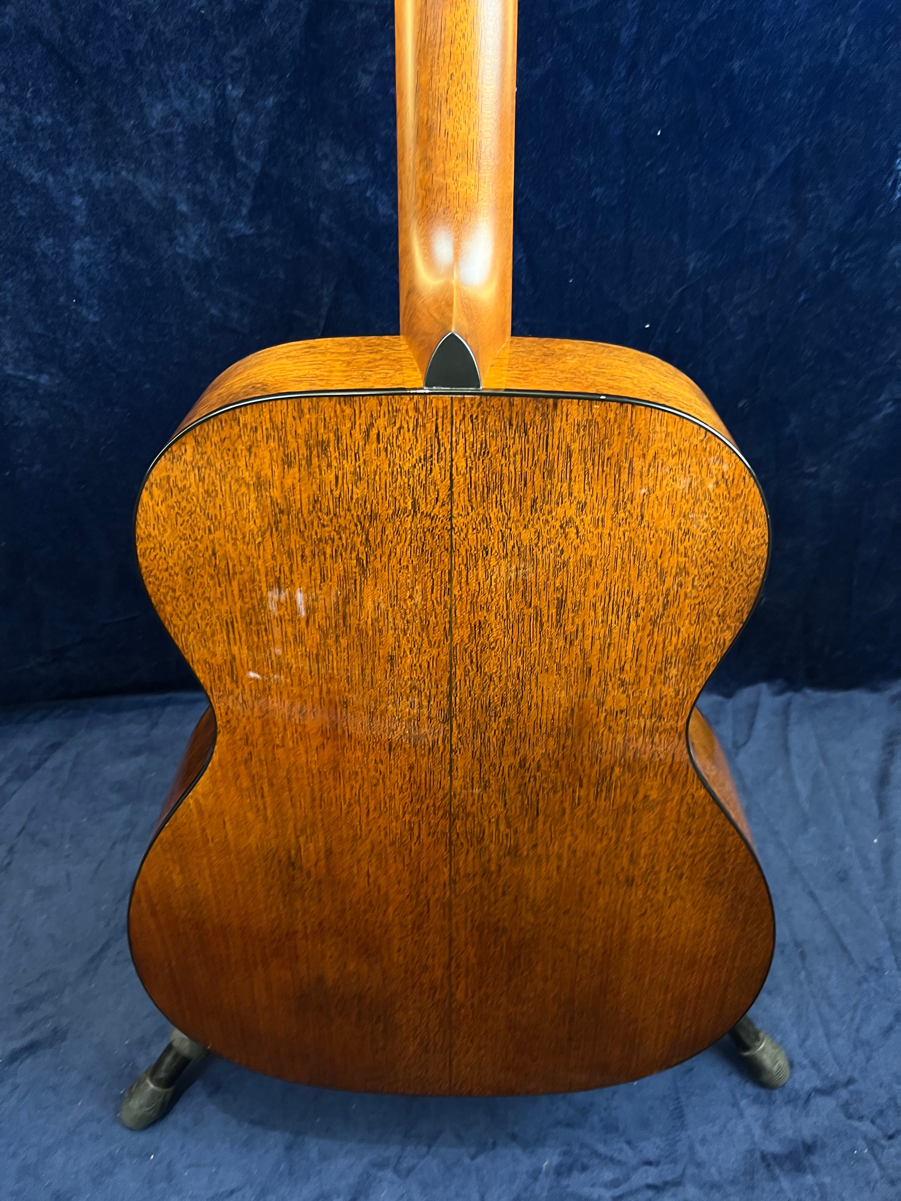 Back view of an acoustic guitar showing a rich brown wood grain finish with a black heel cap where the neck meets the body, standing on a floor stand.