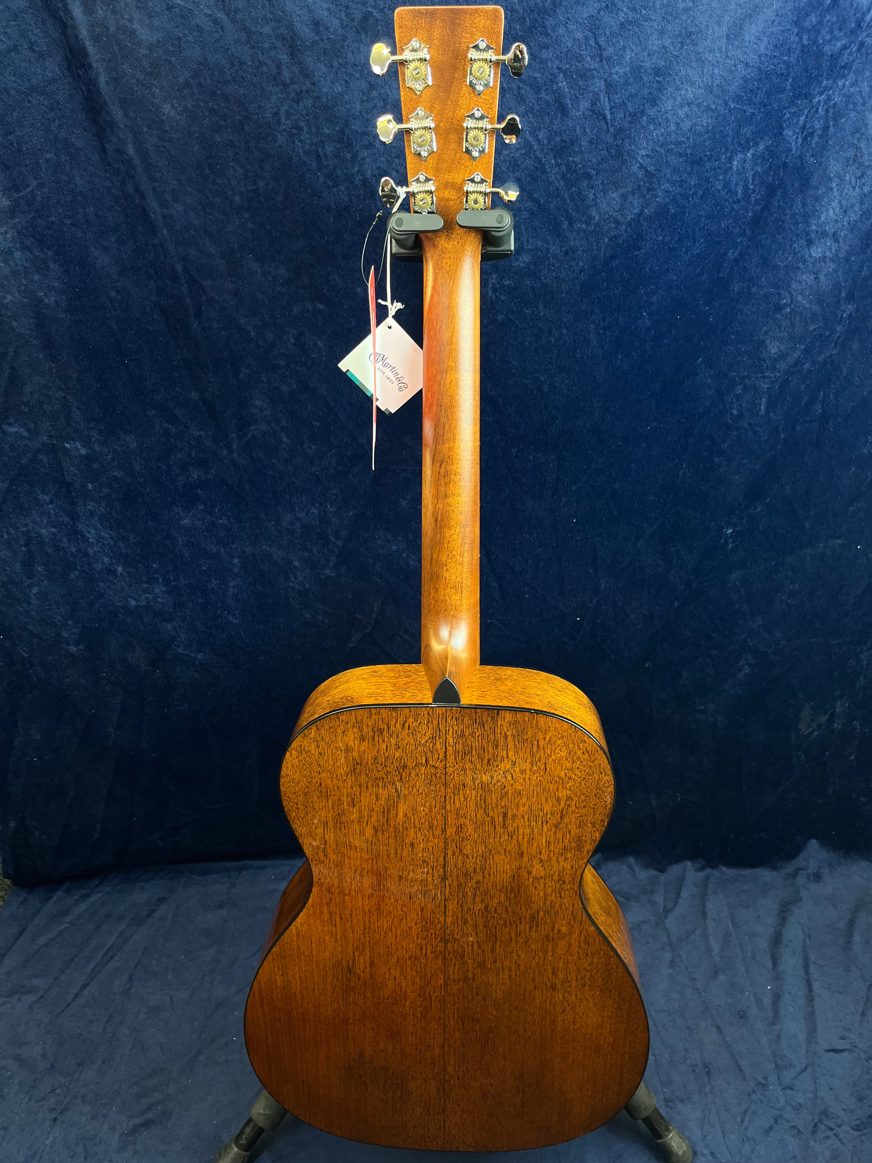 Mahogany acoustic guitar seen from the back, showing its wooden grain, six tuning pegs, and a tag hanging from the headstock.