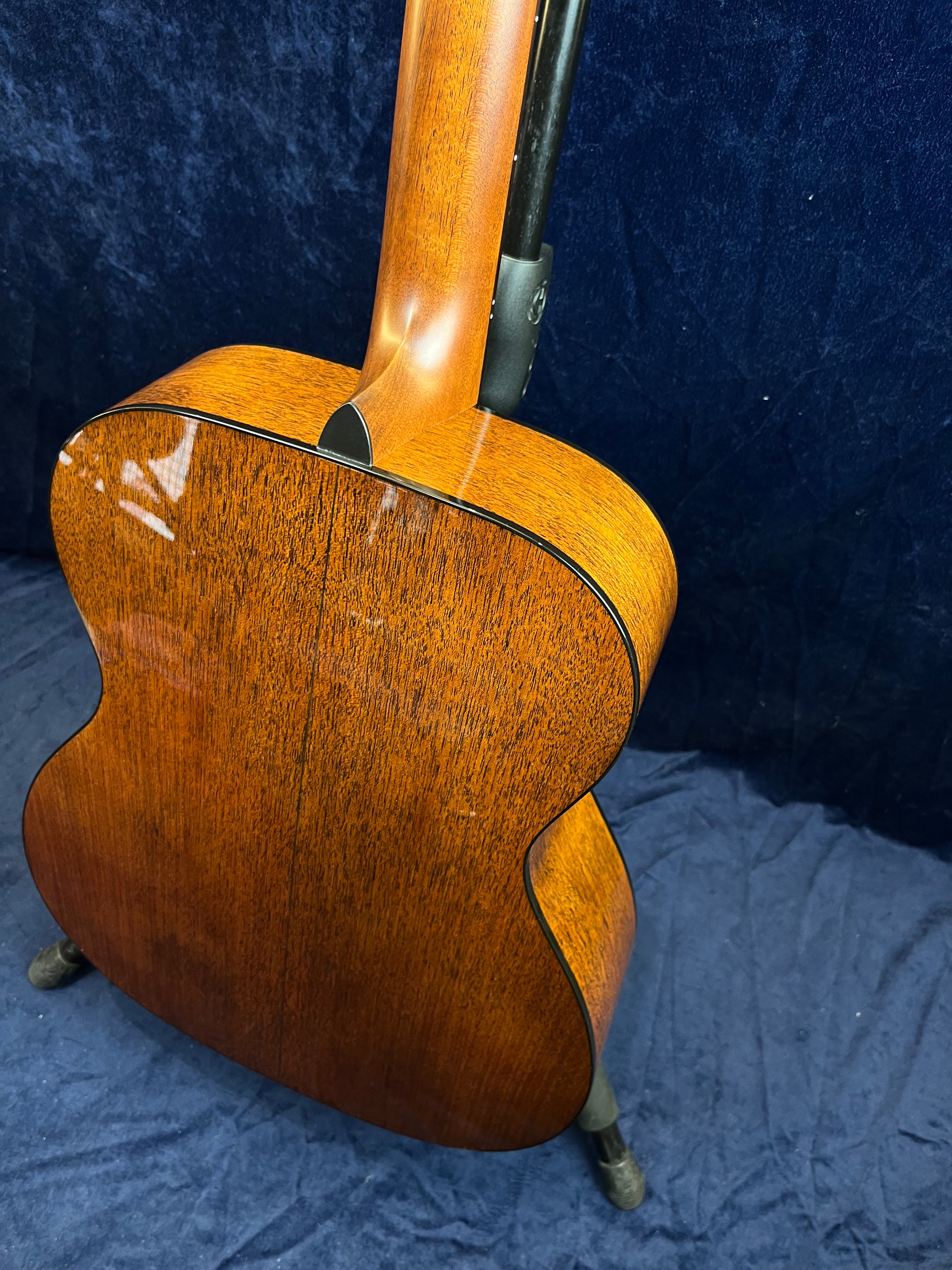 Back and neck of an acoustic guitar made from polished medium-brown wood with a subtle grain pattern, shown on a stand against a blue background.