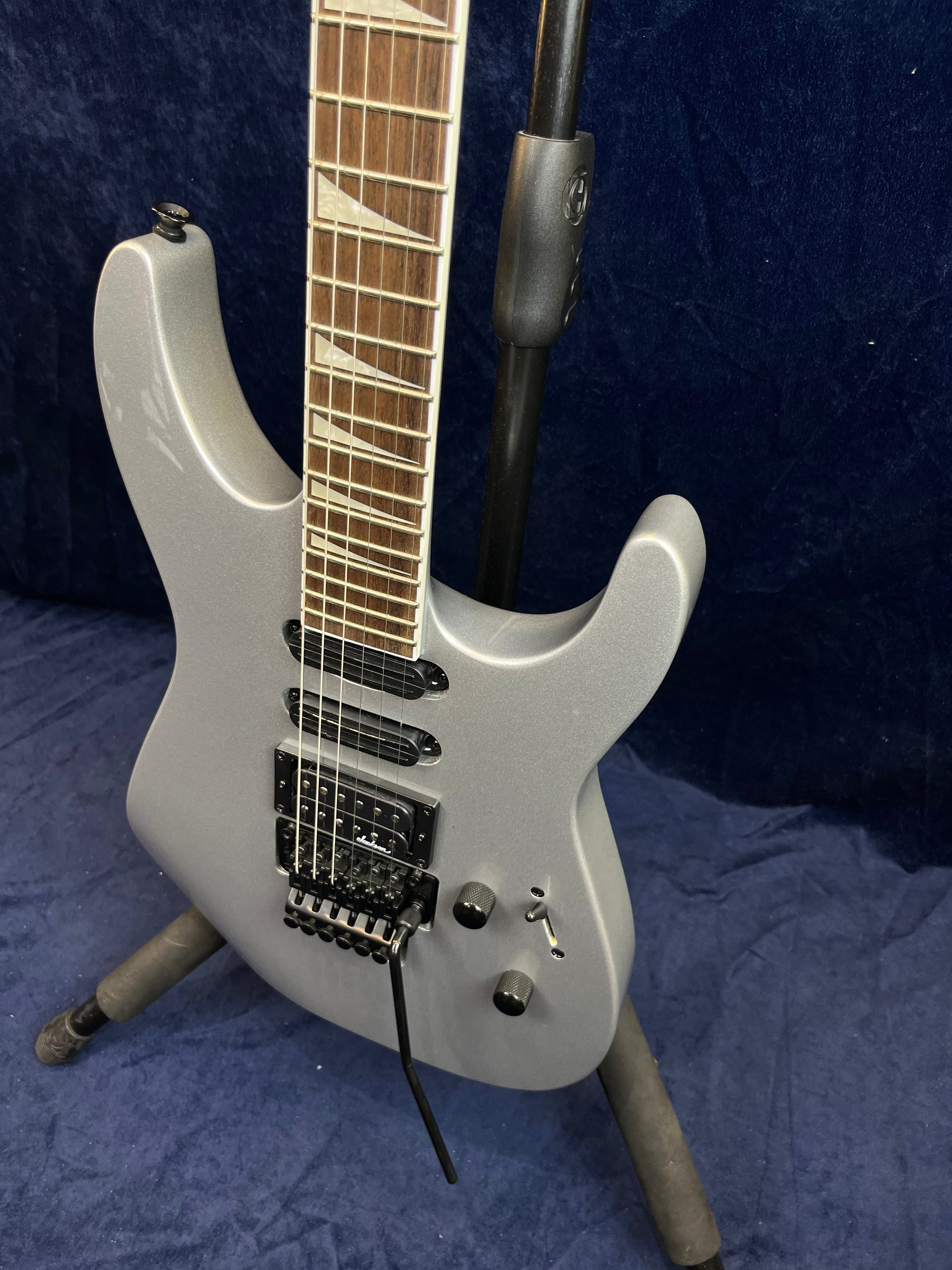 Guitare électrique gris métallisé à corps anguleux, trois micros noirs, vibrato, deux boutons, sélecteur et repères triangulaires sur la touche.