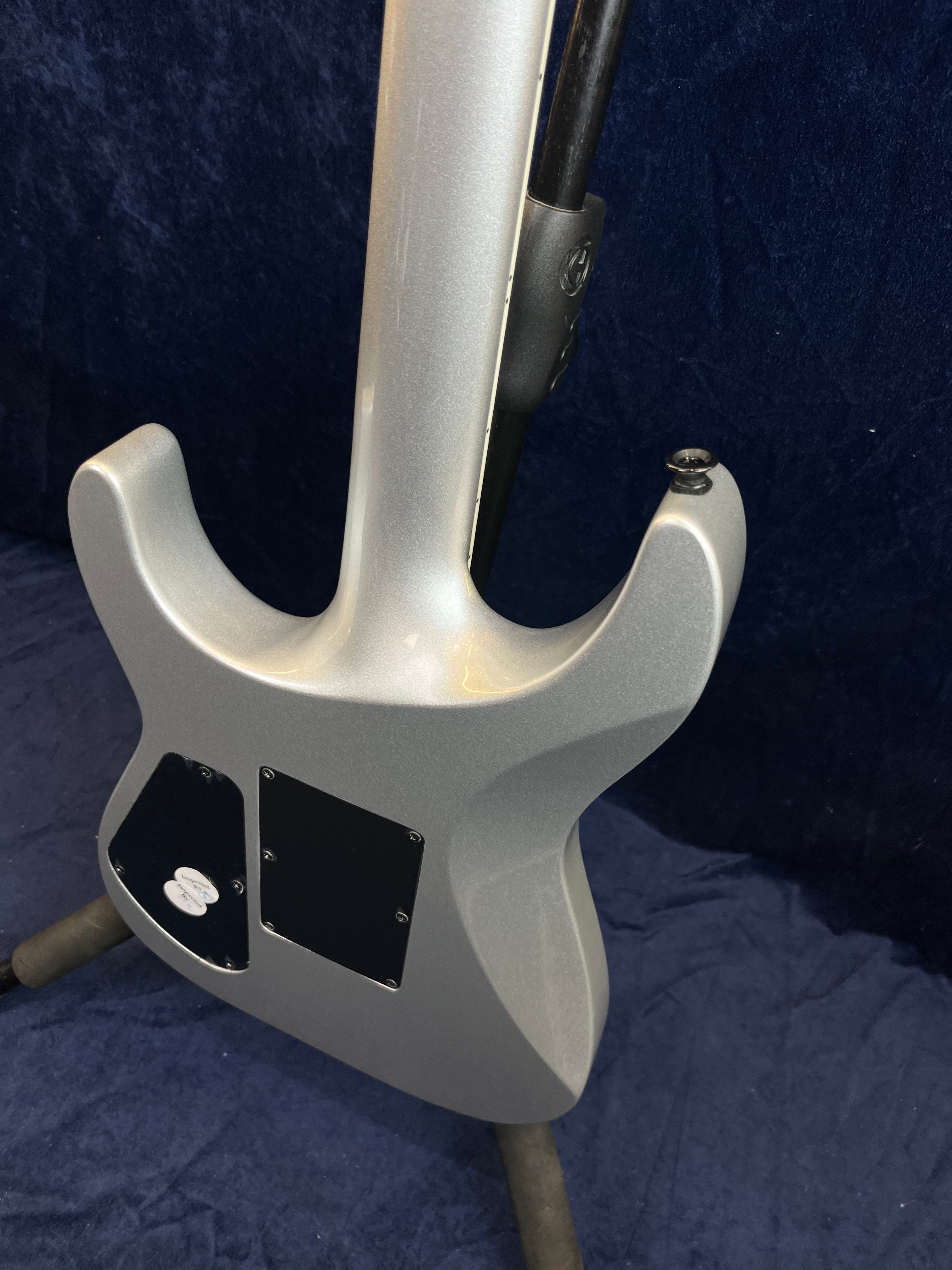 Arrière d’une guitare électrique gris métallisé, avec plaques de contrôle noires et attaches pour sangle, posée sur un support contre un fond bleu.