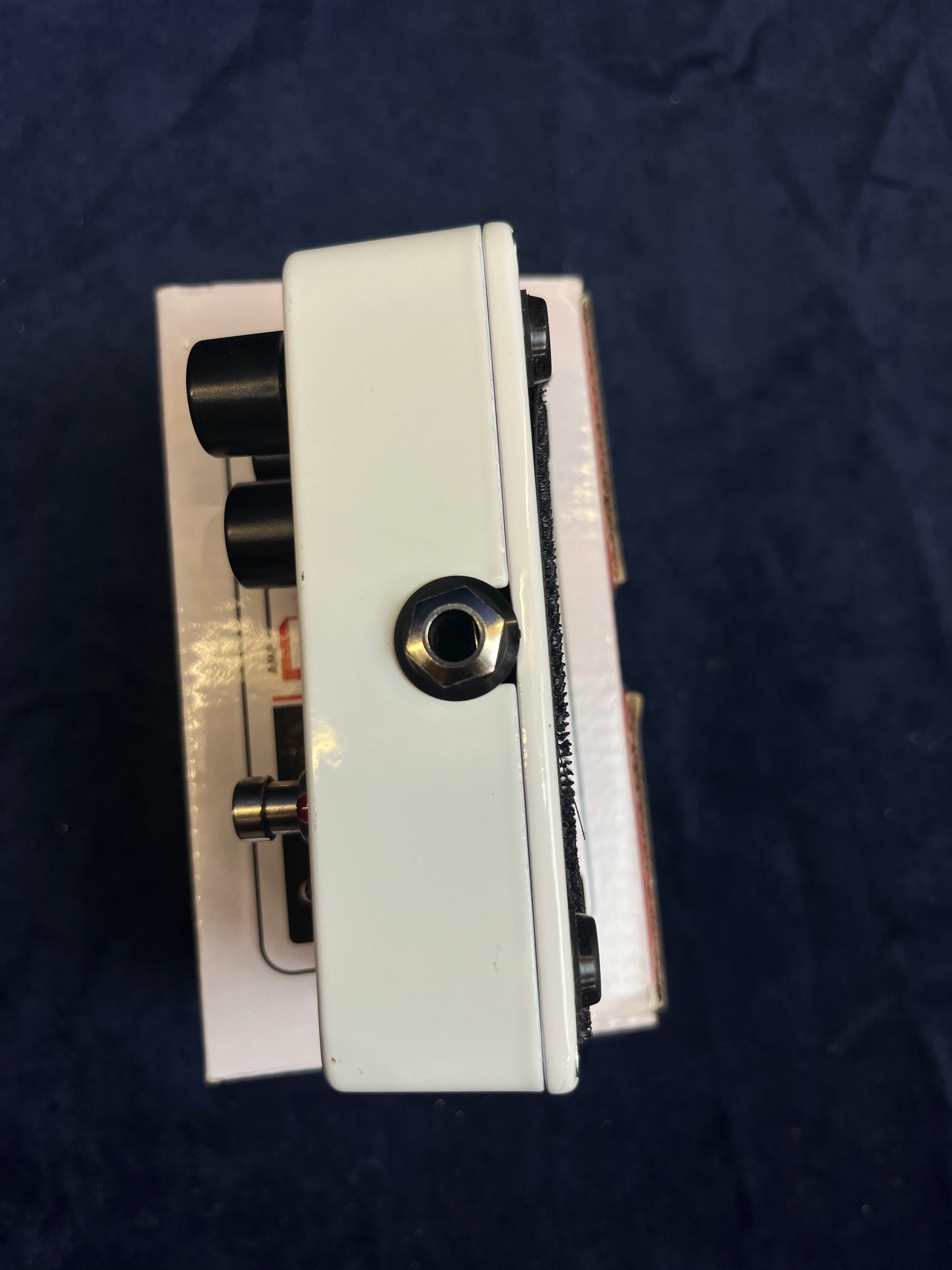White FX pedal with three black knobs on the side, a metal footswitch, and a single input jack, resting on a white box against a dark background.