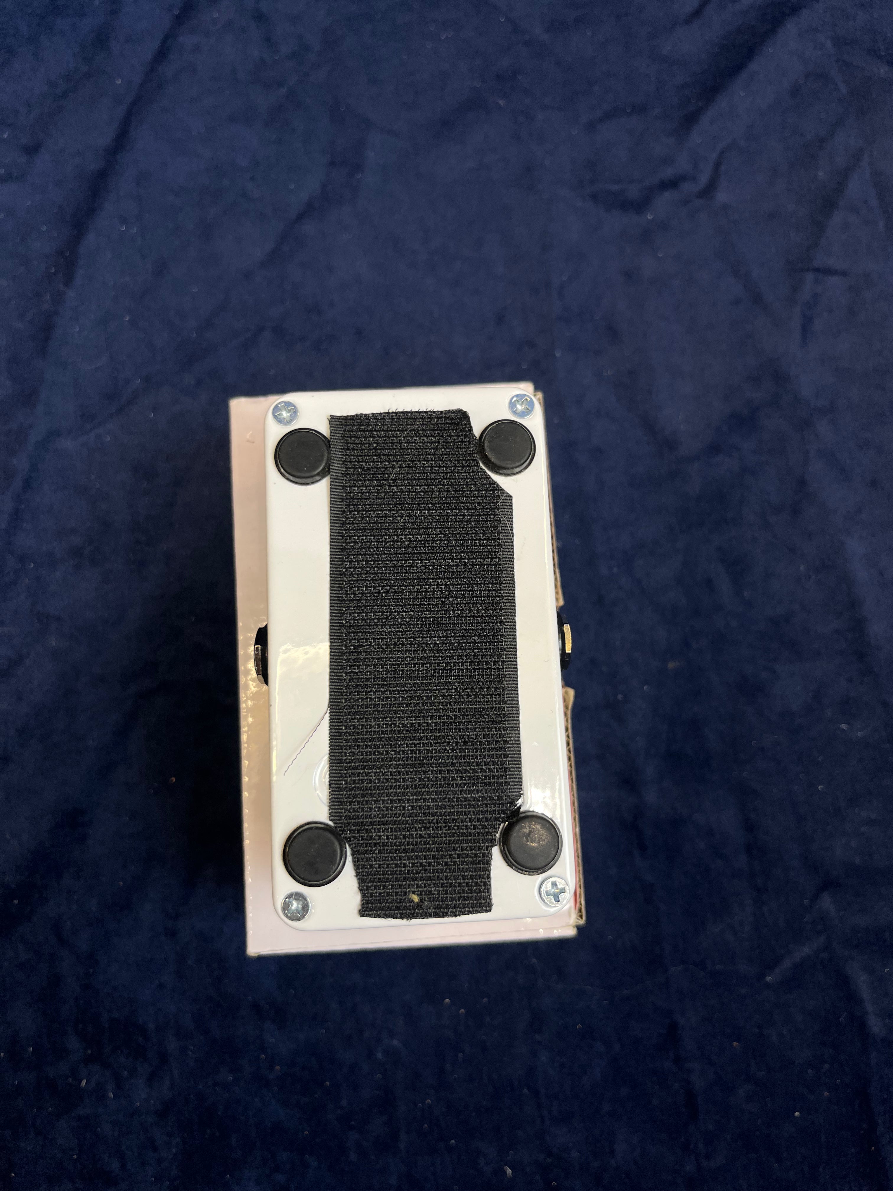 Bottom of an FX pedal showing four rubber feet, four screws, and a strip of black Velcro attached for pedalboard mounting, on a dark blue surface.