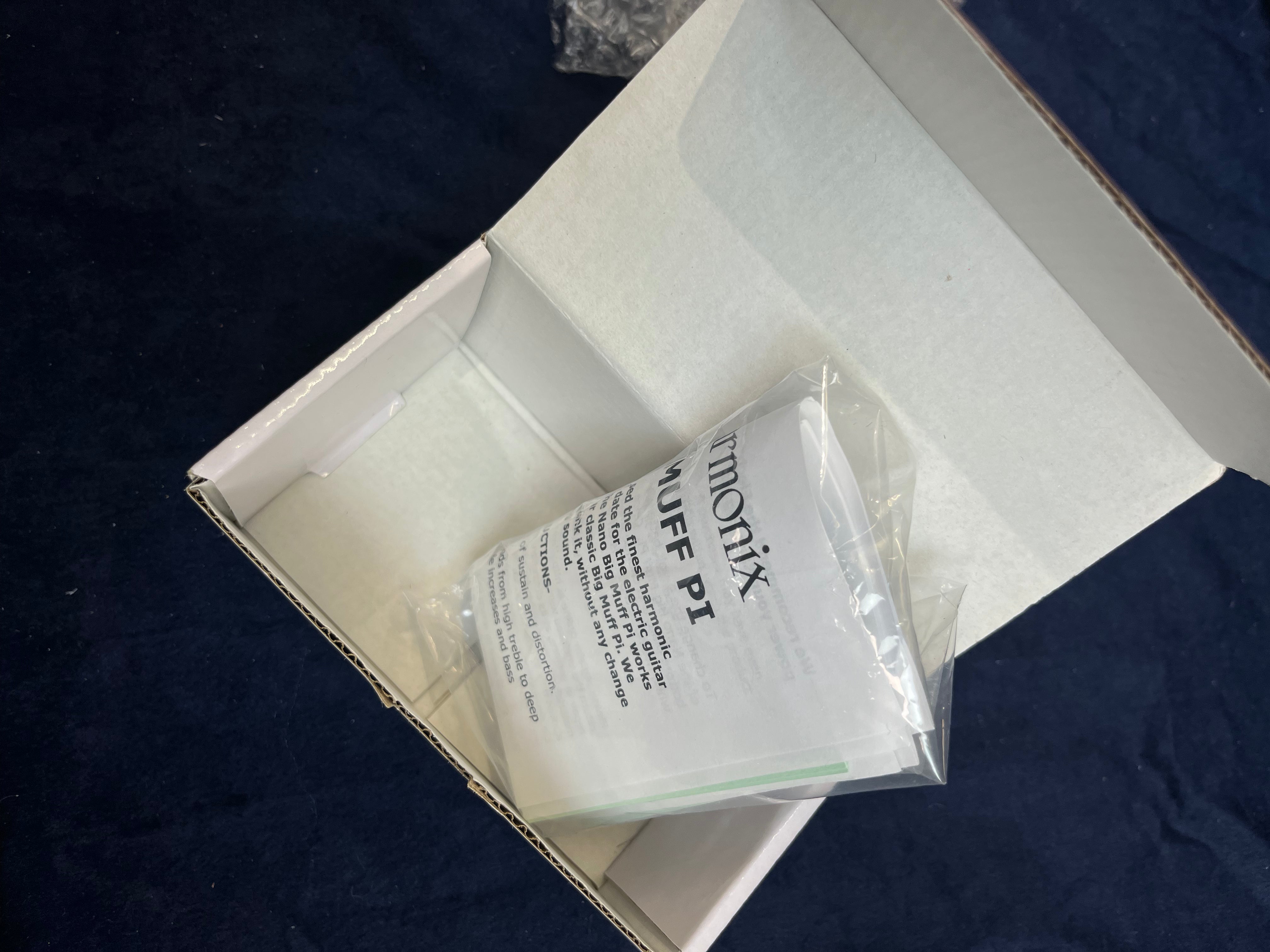 Open box on dark fabric, containing an instruction manual for an FX pedal labeled "Harmonix MUFF PI" sealed in a clear plastic bag.