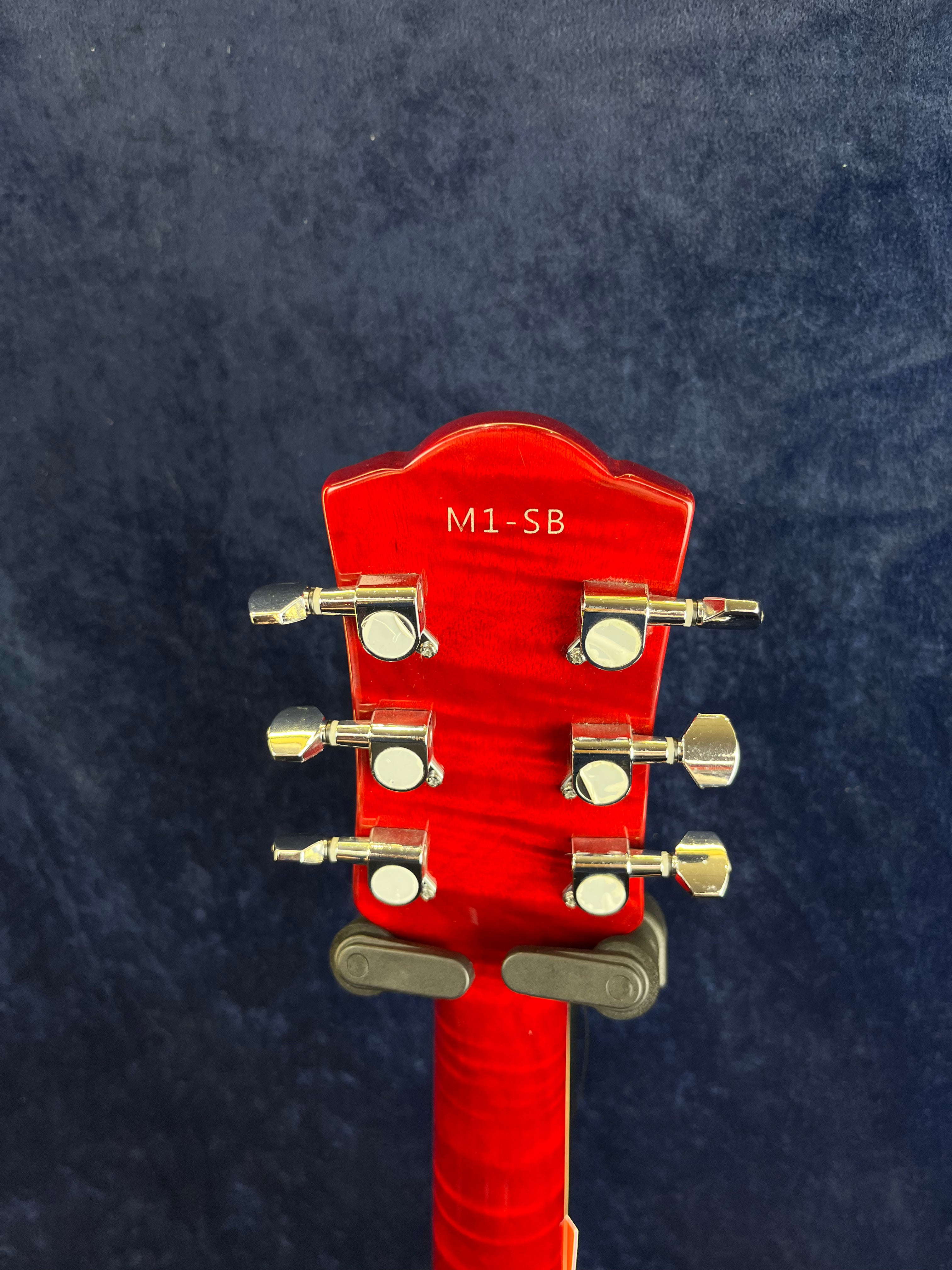 Red electric guitar headstock with six silver tuning pegs and “M1-SB” printed in white text on the back, set against a dark blue background.