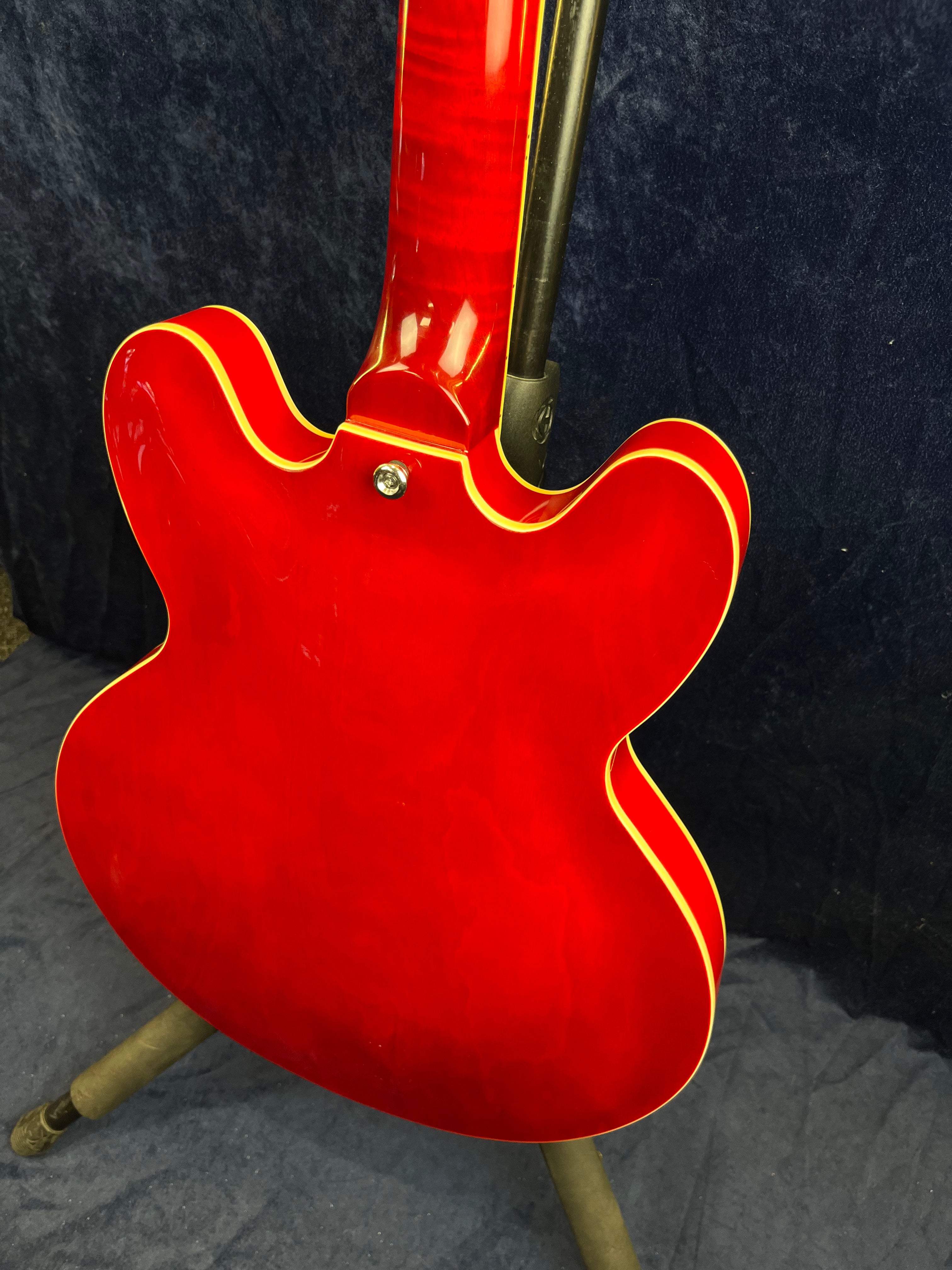 Bright red semi-hollow electric guitar shown from the back, with cream-colored binding around the edges, resting on a stand against a dark background.