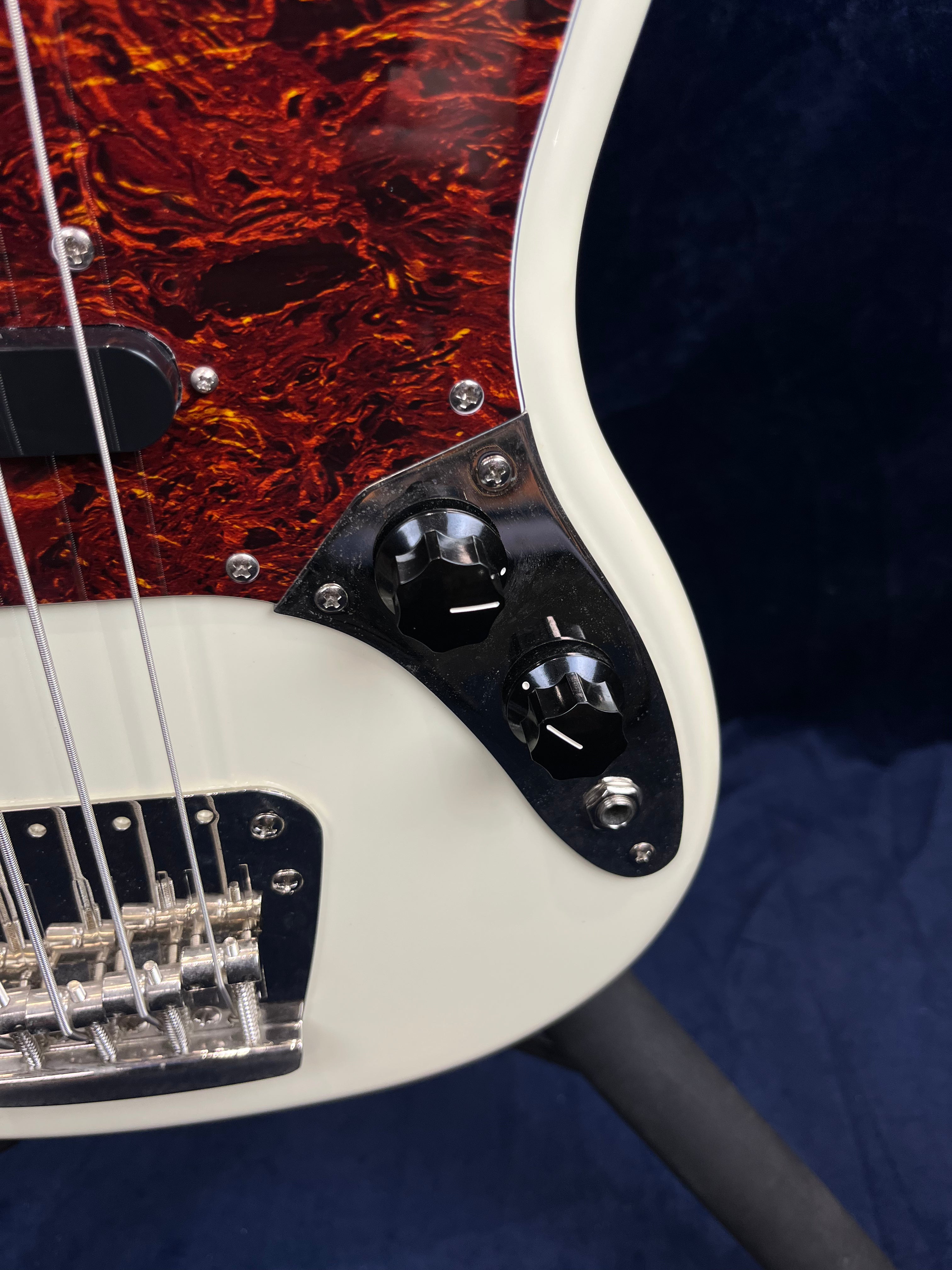 Close-up of a white bass guitar body showing two black control knobs, a metal bridge, and a tortoiseshell pickguard with metal screws.