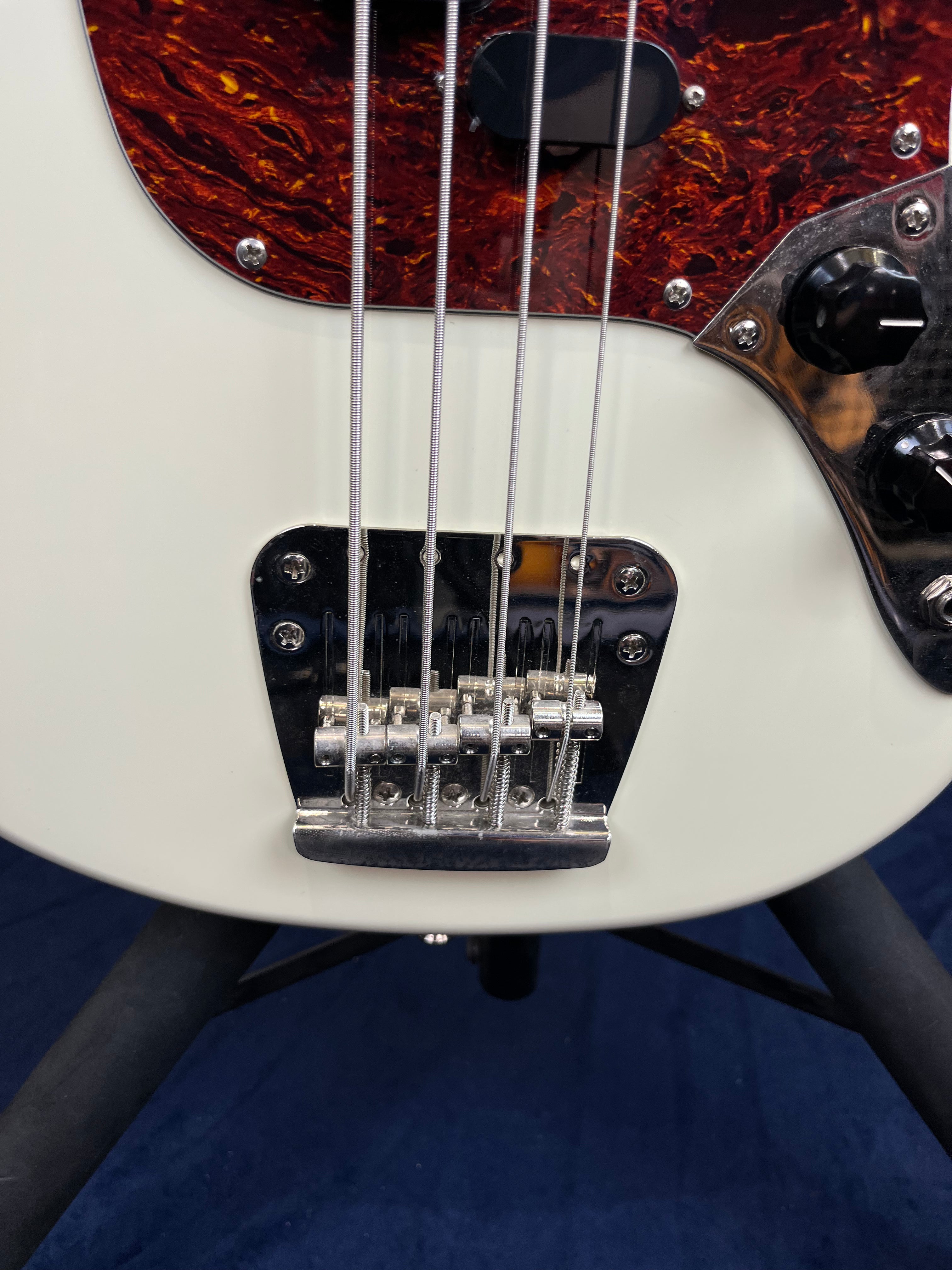 White bass guitar body with four metal strings, a chrome bridge, tortoiseshell pickguard, and black control knobs visible on a stand.