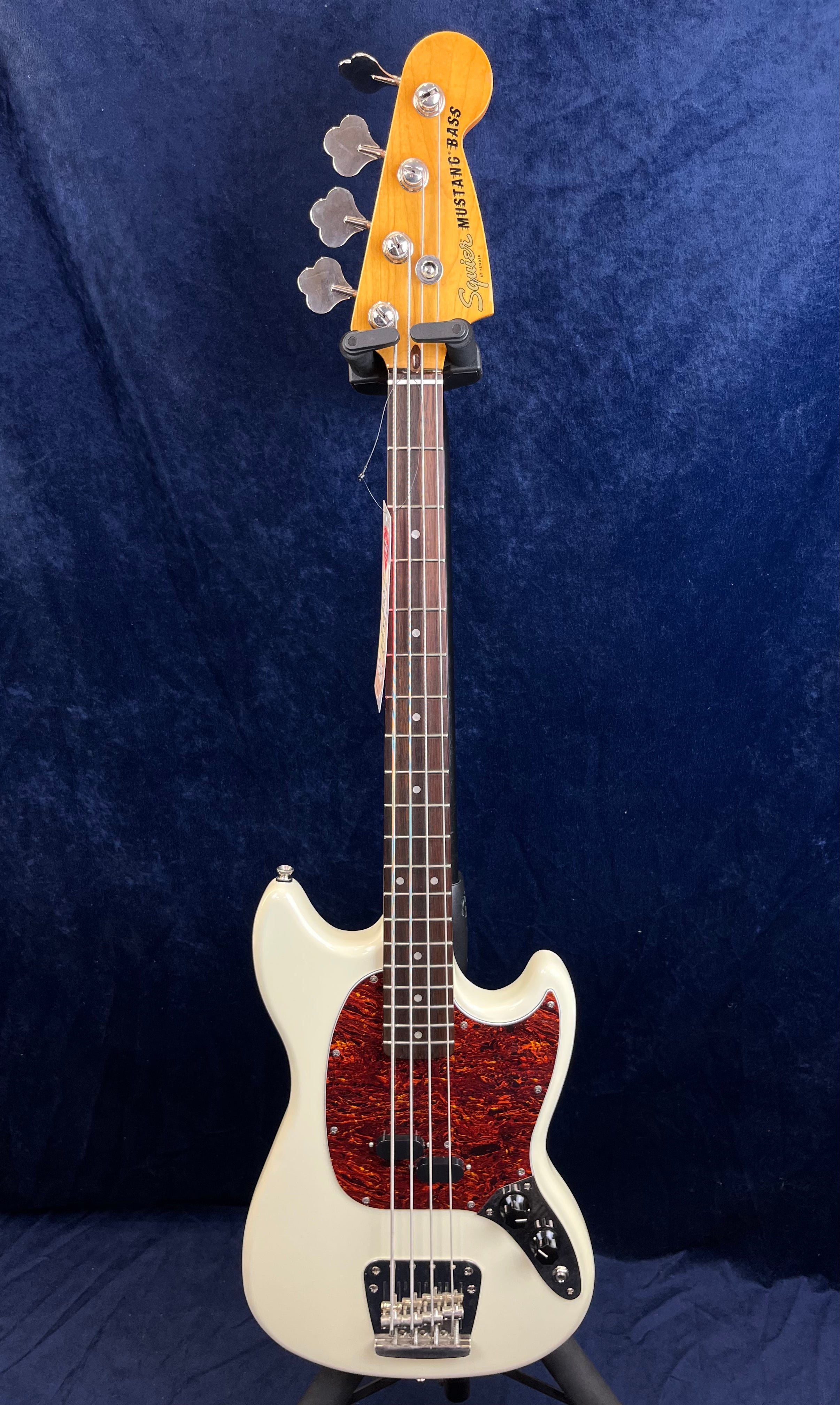 Squier Mustang Bass guitar with an off-white body, tortoiseshell pickguard, rosewood fingerboard, and chrome hardware standing upright.