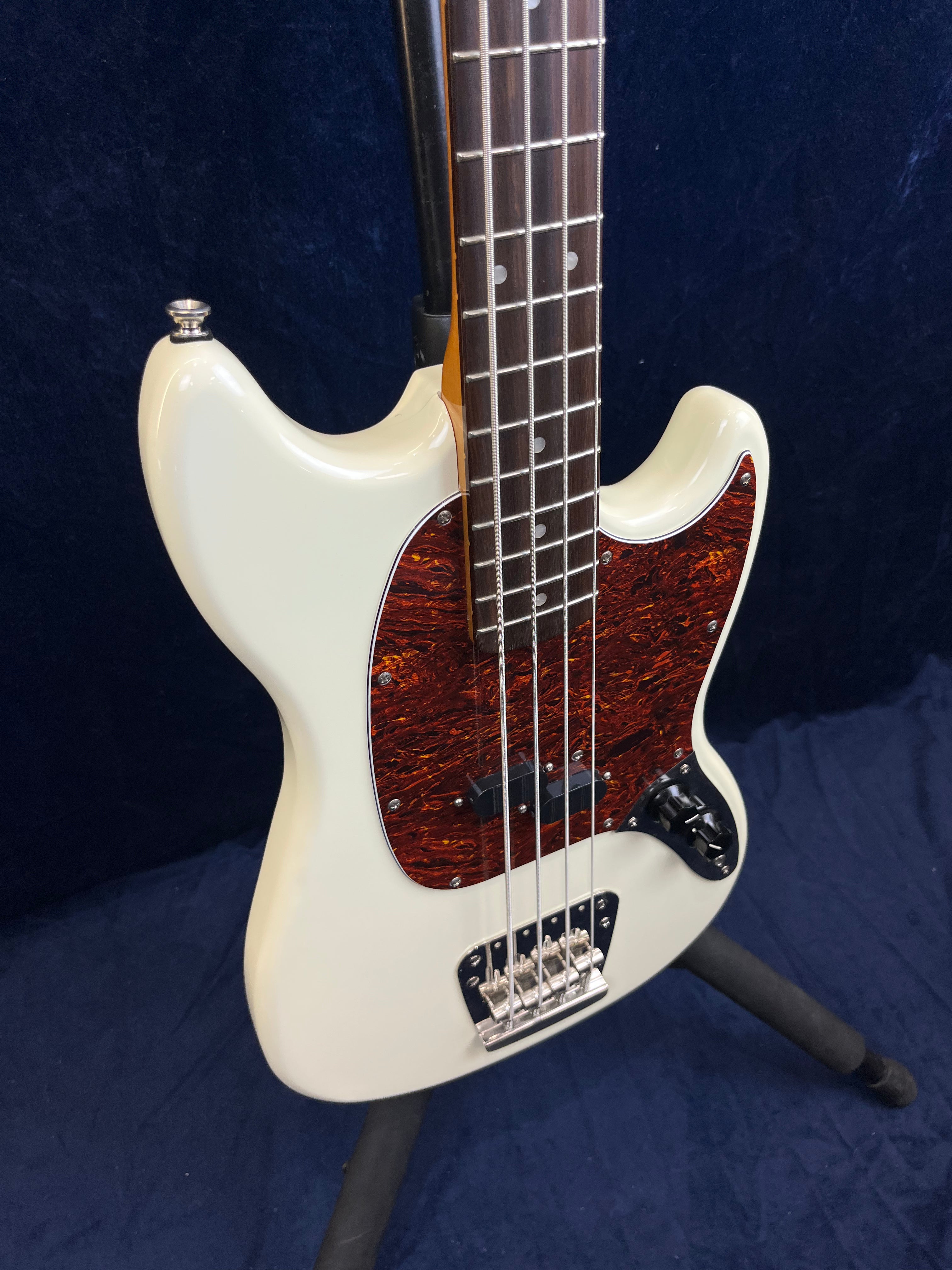 White bass guitar with a dark brown tortoiseshell pickguard, four strings, and silver hardware, displayed upright on a stand.