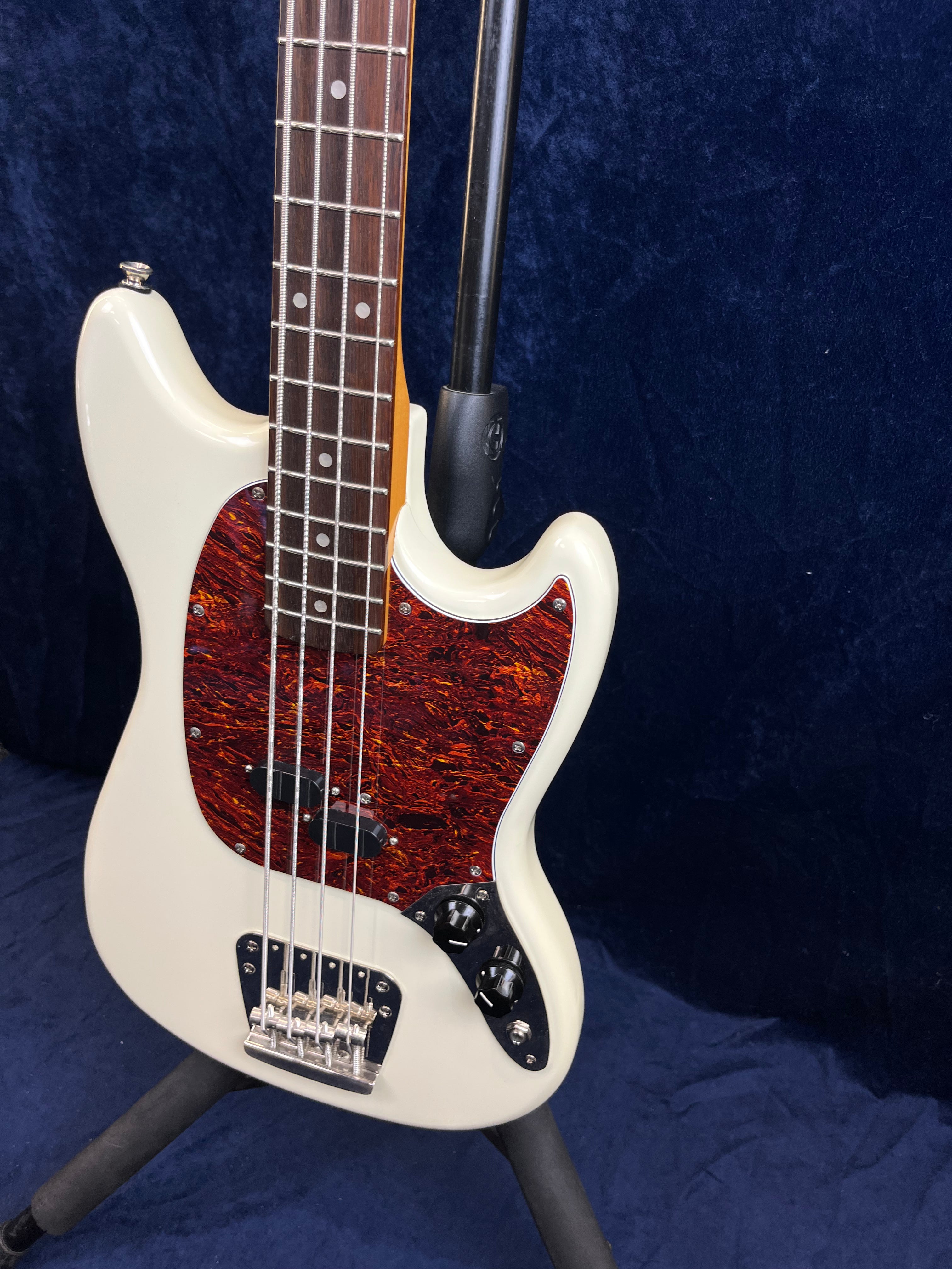 White bass guitar with a brown tortoiseshell pickguard, chrome bridge, and black control knobs, shown on a stand against a dark blue background.
