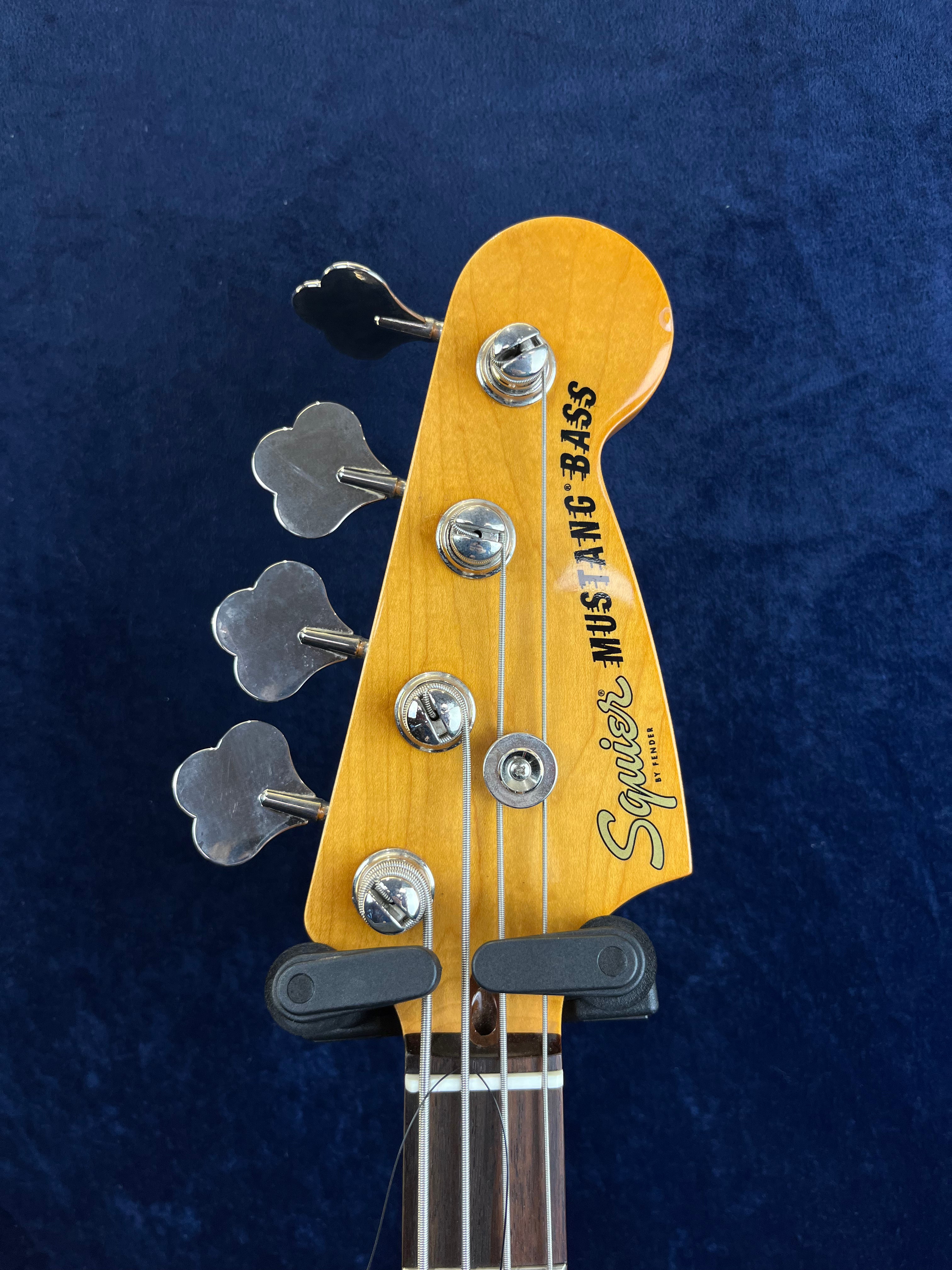 Squier bass guitar headstock with four tuning pegs, chrome hardware, and "JAZZ BASS" label on a natural wood finish against a dark blue background.