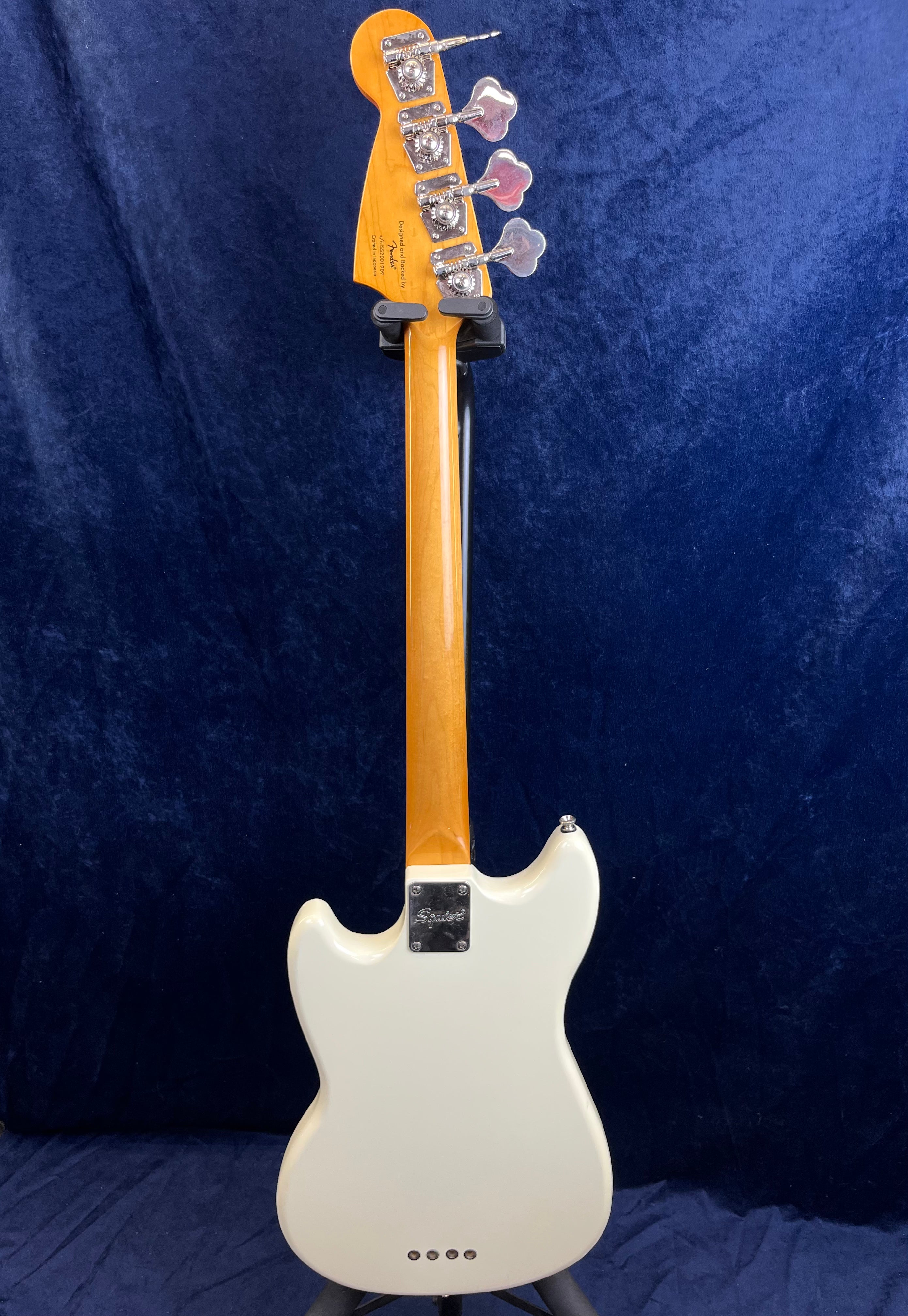 White Squier bass guitar shown from the back, with a light wood neck and silver tuning pegs against a dark blue background.