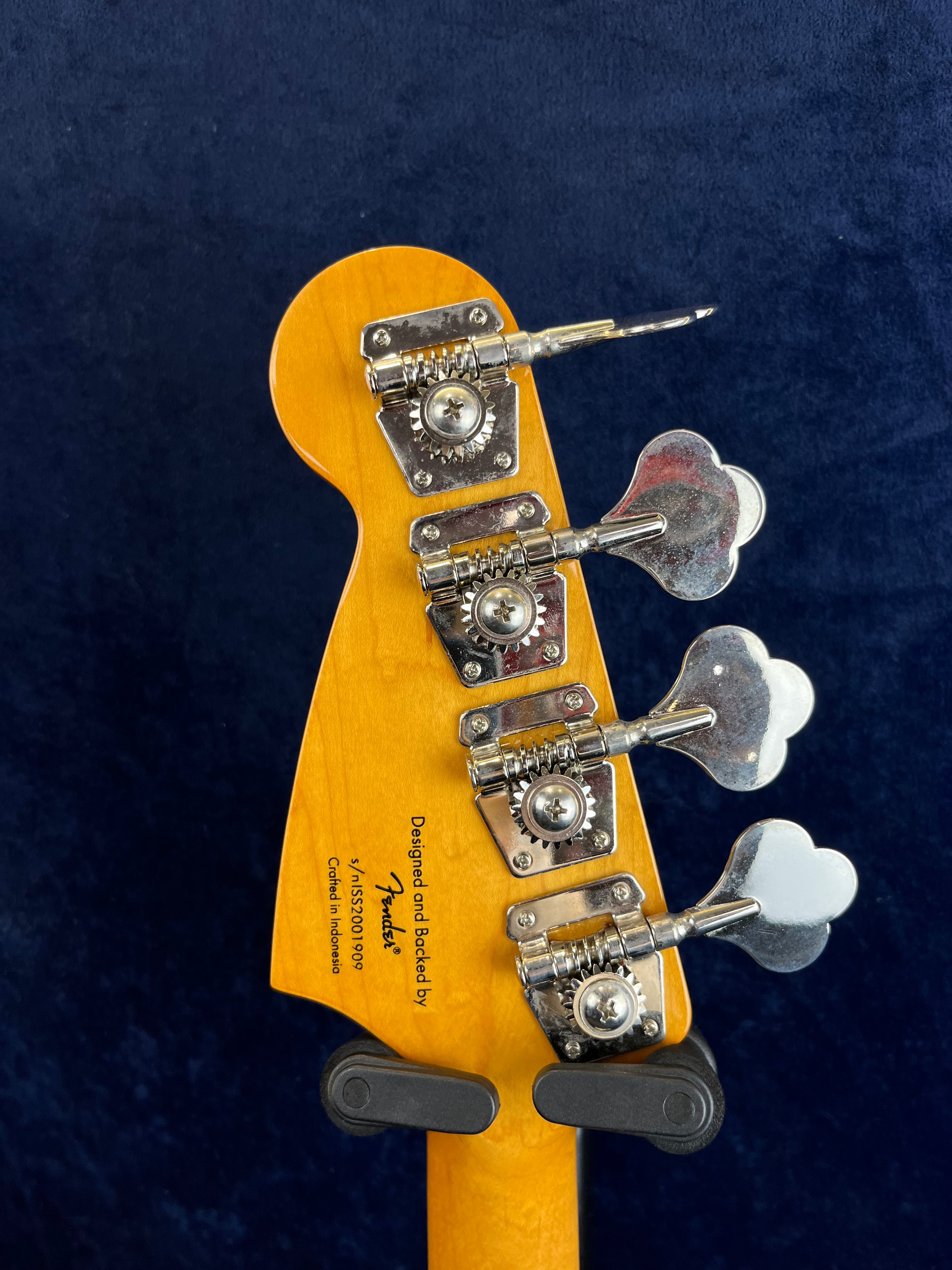 Four large chrome tuning machines on a light wood Fender bass guitar headstock, black text labeling “Designed and Backed by Fender” is visible.
