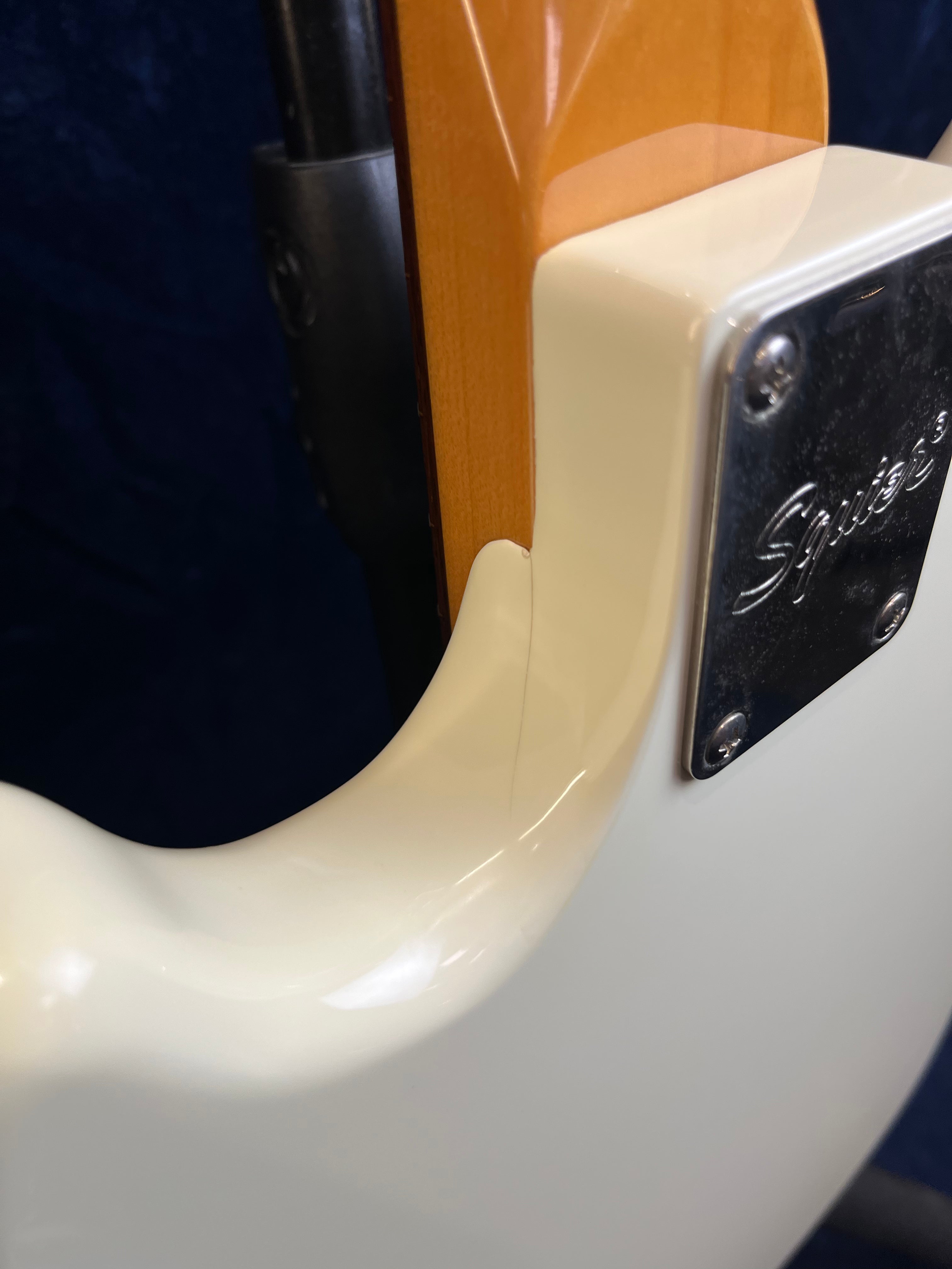 Closeup of a cream-colored Squier bass guitar’s neck joint, showing the neck plate, part of the body, and glossy wood finish.