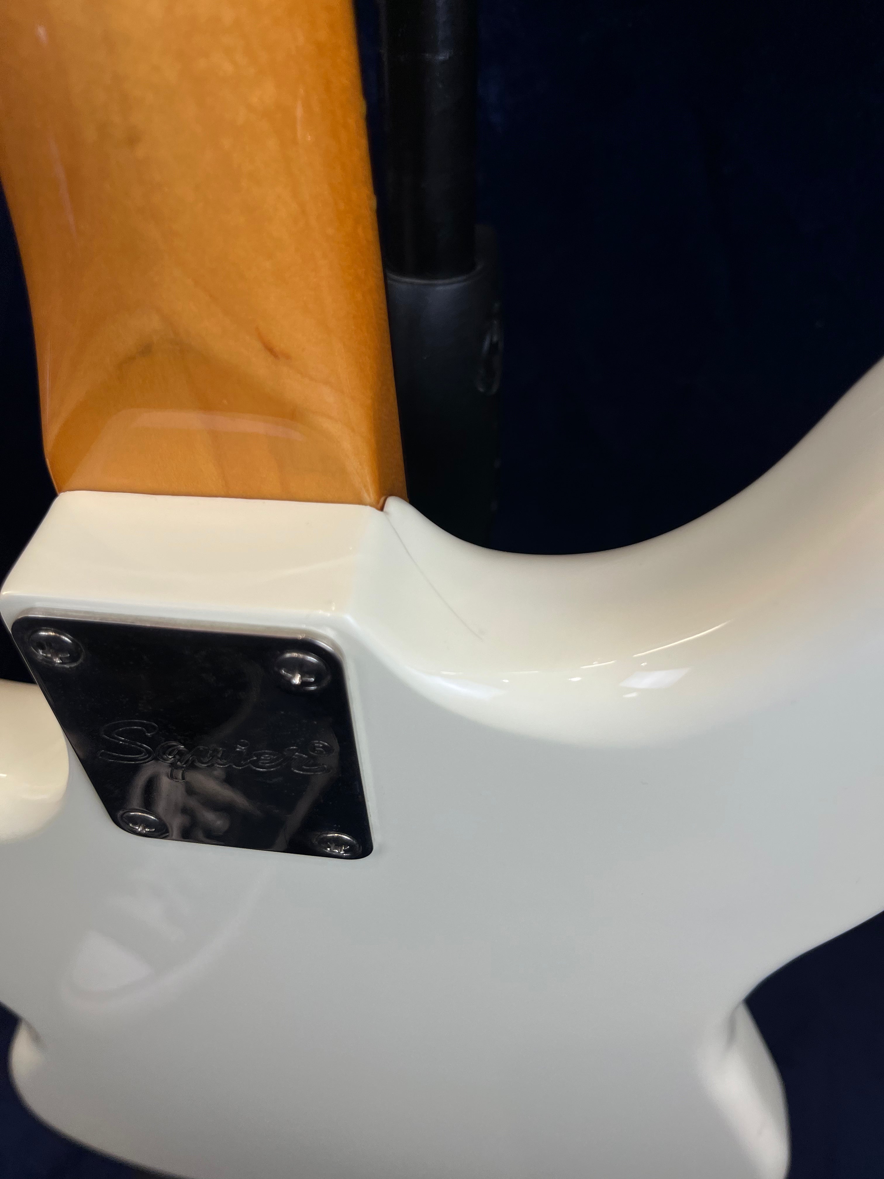 Close-up of a Squier bass guitar’s neck joint showing a white body, metal neck plate with four screws, and a light brown wooden neck.