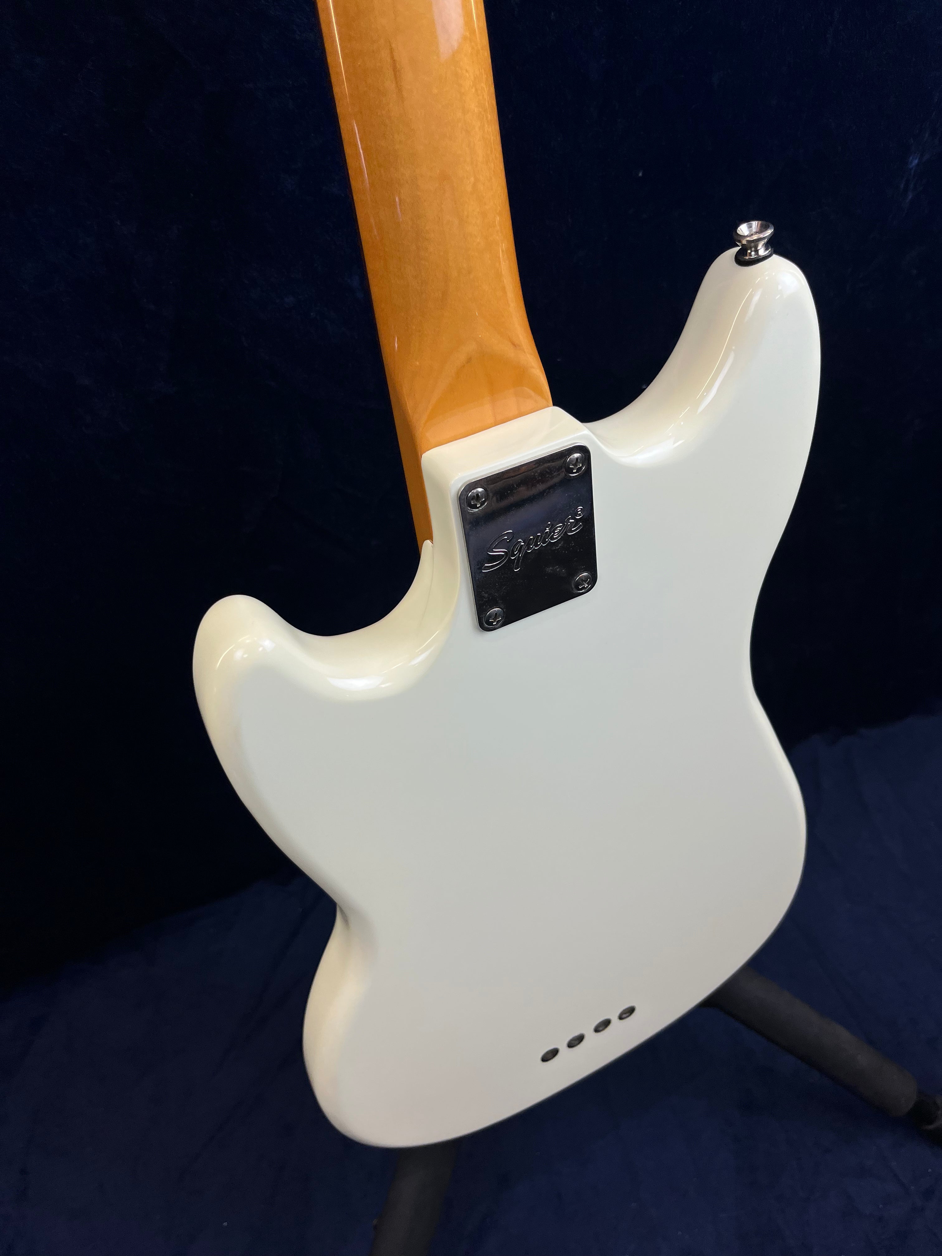 White Squier bass guitar shown from the back, highlighting its glossy finish, neck joint plate, and four body holes near the bottom.