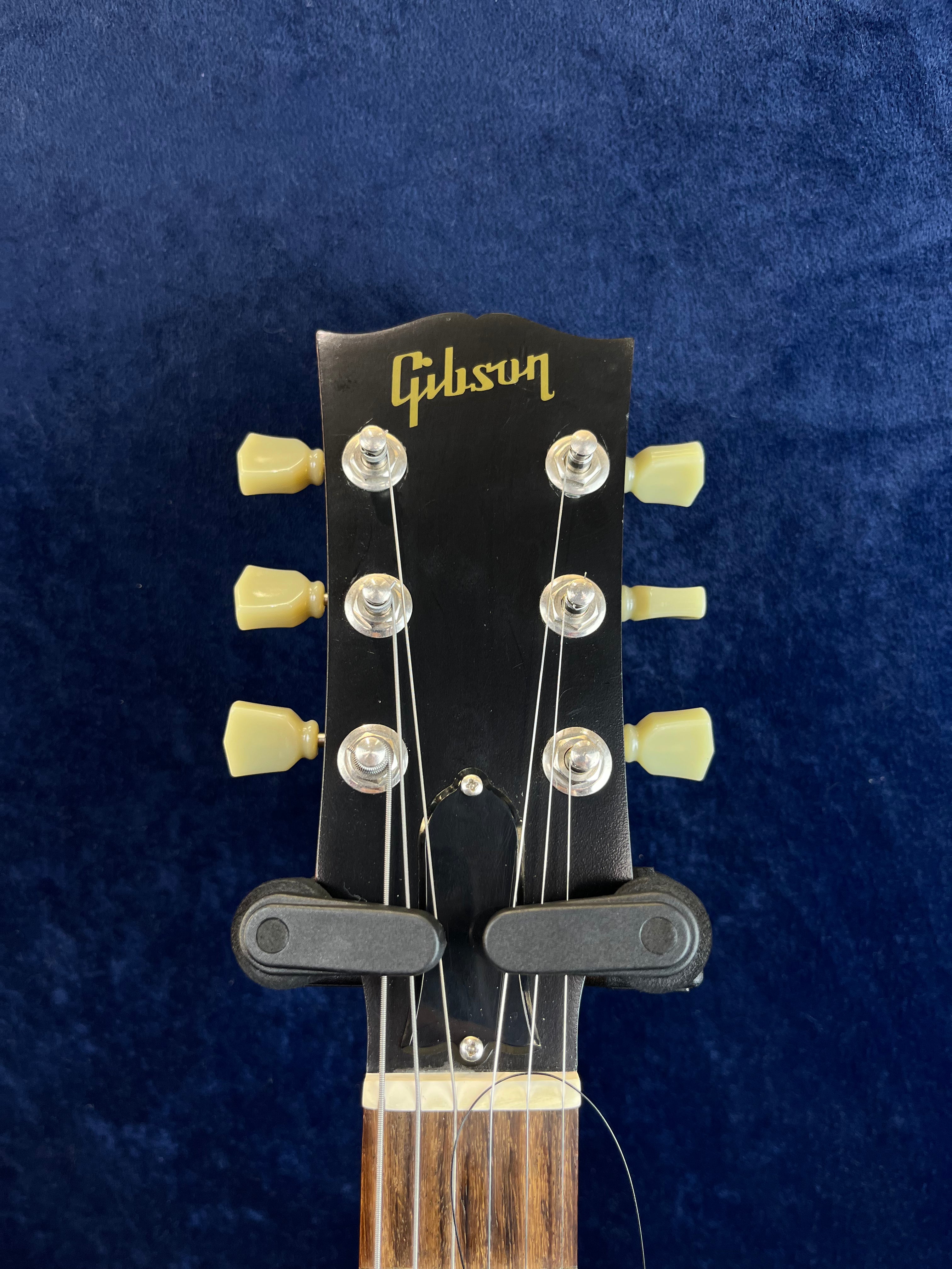 Gibson electric guitar headstock with six cream tuning pegs on a dark background, some loose guitar strings visible, wood fretboard at bottom.